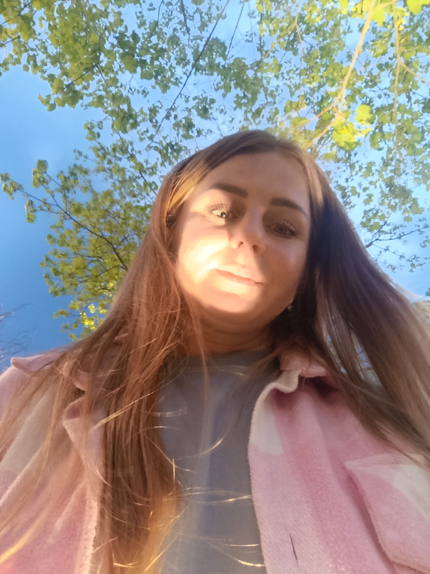 A woman with long, light brown hair is looking down at the camera. She is wearing a gray shirt and a pink and white flannel. The background is a blue sky with green leaves and branches from a tree. The location is in the city of Unknown, Unknown.