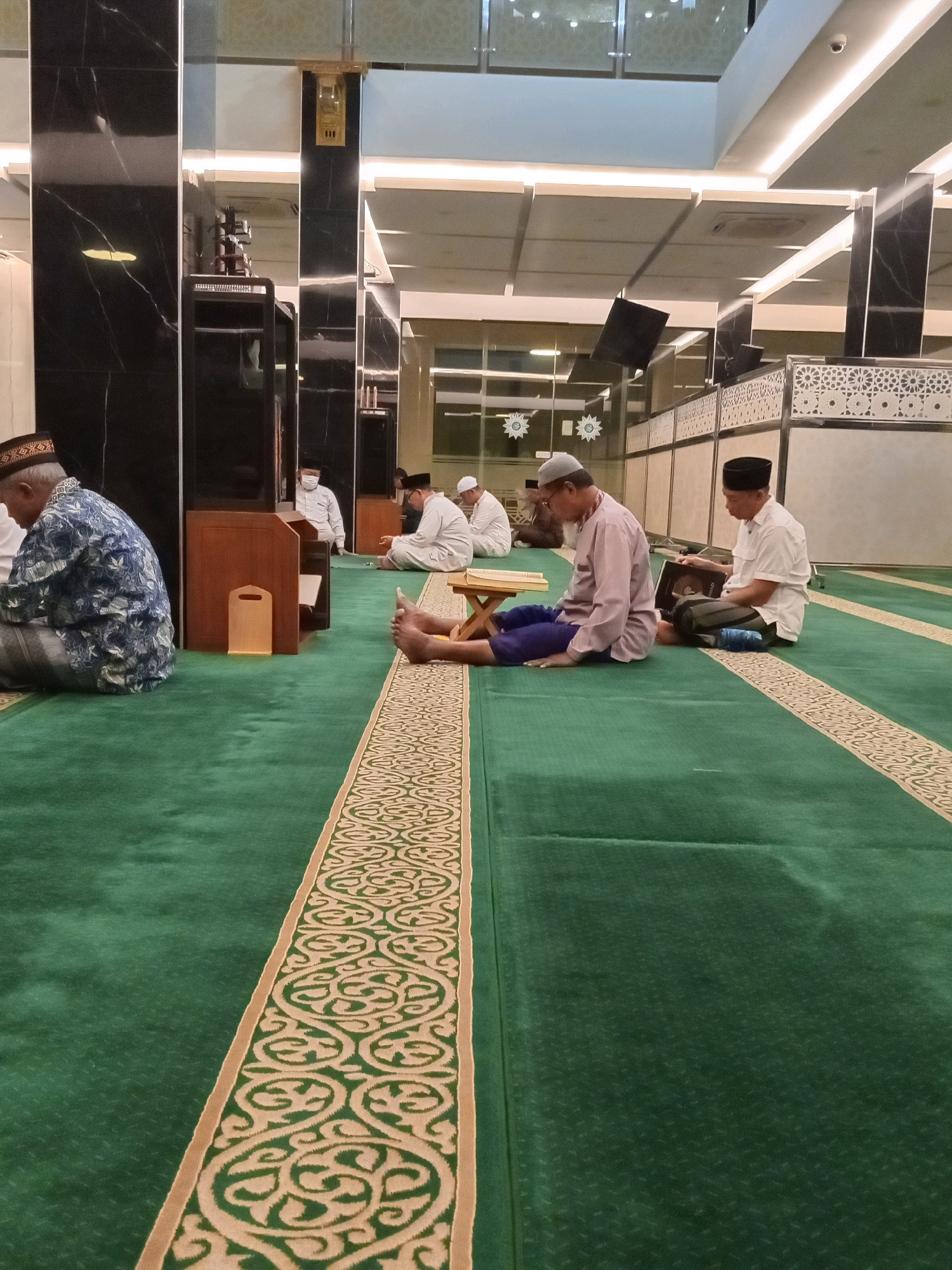 In the city of Randuagung, Indonesia, an indoor scene unfolds within a mosque characterized by a predominantly green carpet with a beige and tan patterned border. Several men are present, engaged in prayer or contemplation. In the foreground, on the left, a man wearing a patterned blue shirt and a dark cap with a textured brim is seated on the carpet. To his right, closer to the center, another man in a light purple shirt and blue pants is seated, with a small wooden stand holding an open book in front of him. Further to the right, a man in a white shirt and dark pants is seated, also reading from an open book. In the background, other individuals are visible, including one in a white shirt and cap standing near a wooden structure. The mosque's interior features dark, reflective pillars, bright overhead lighting, and patterned wall panels. The overall atmosphere suggests a moment of religious observance.
