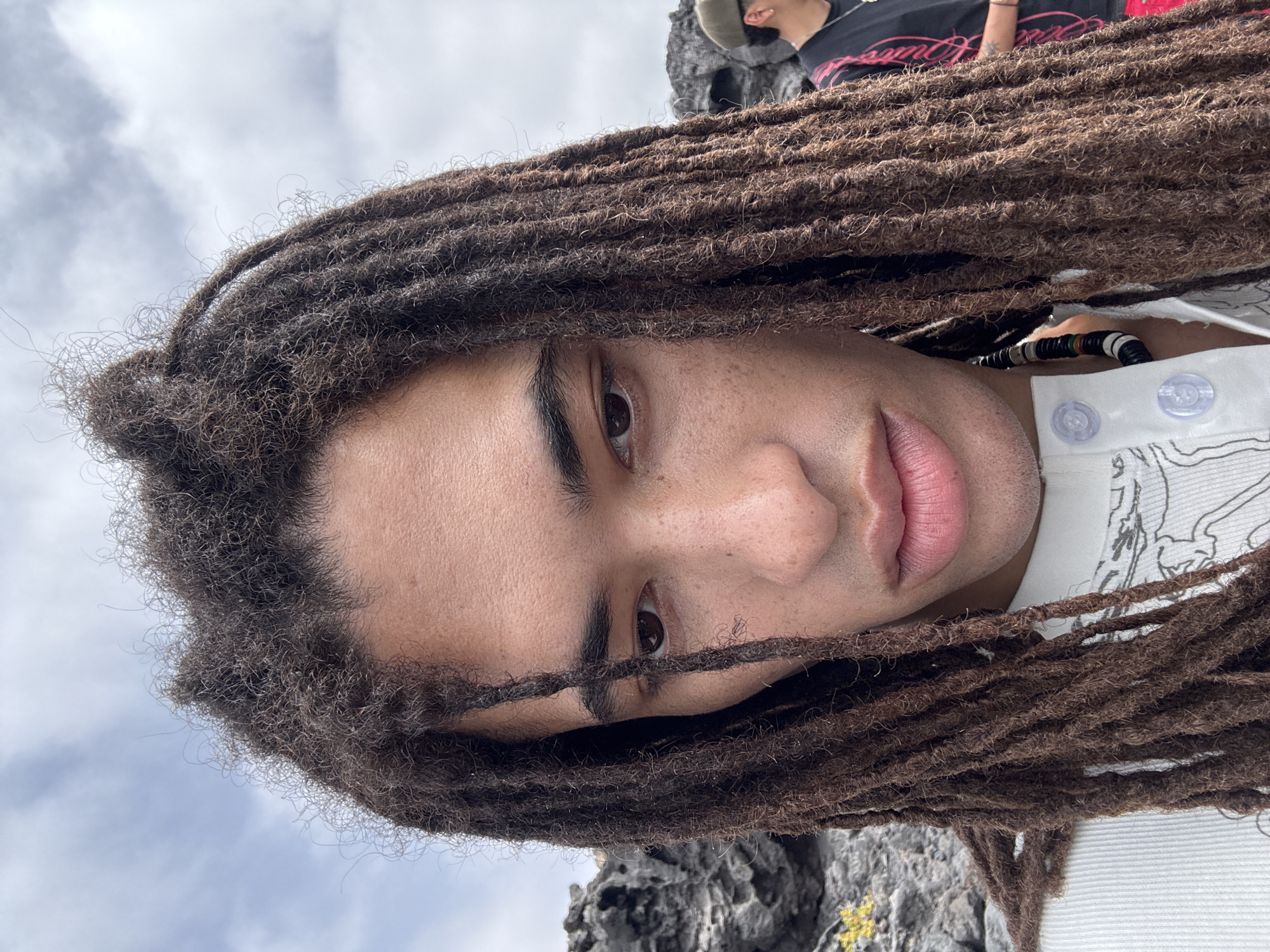 A portrait-style photograph features a person with dark, tightly coiled dreadlocks framing their face. The individual's skin tone is light brown, with visible freckles. They are wearing a white shirt with visible buttons.
In the background, there are indistinct figures and a rocky landscape. The location is speculated to be in the city of Tinajo, Spain.