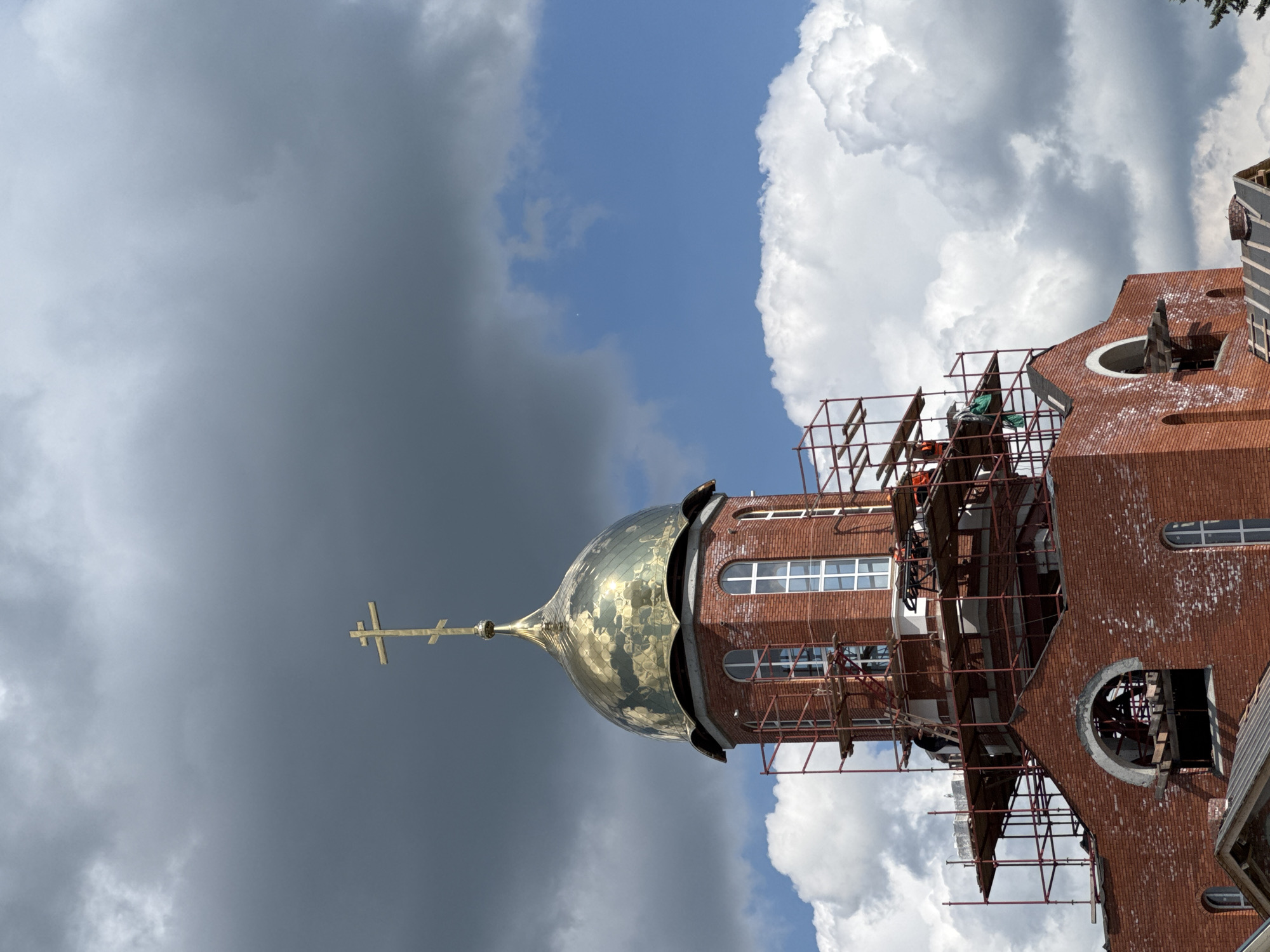 A close-up, low-angle, vertical shot shows a partially constructed red brick church topped with a golden dome and a cross, set against a cloudy sky. Scaffolding surrounds the upper portion of the church, with several workers visible in orange vests performing construction tasks. The dome is elaborately shaped and reflects the sky. The sky itself is a dramatic mix of dark, ominous clouds on the left and bright, fluffy white clouds on the right, with patches of blue visible. The church features arched windows with white frames. Some parts of the brickwork appear weathered or incomplete.
