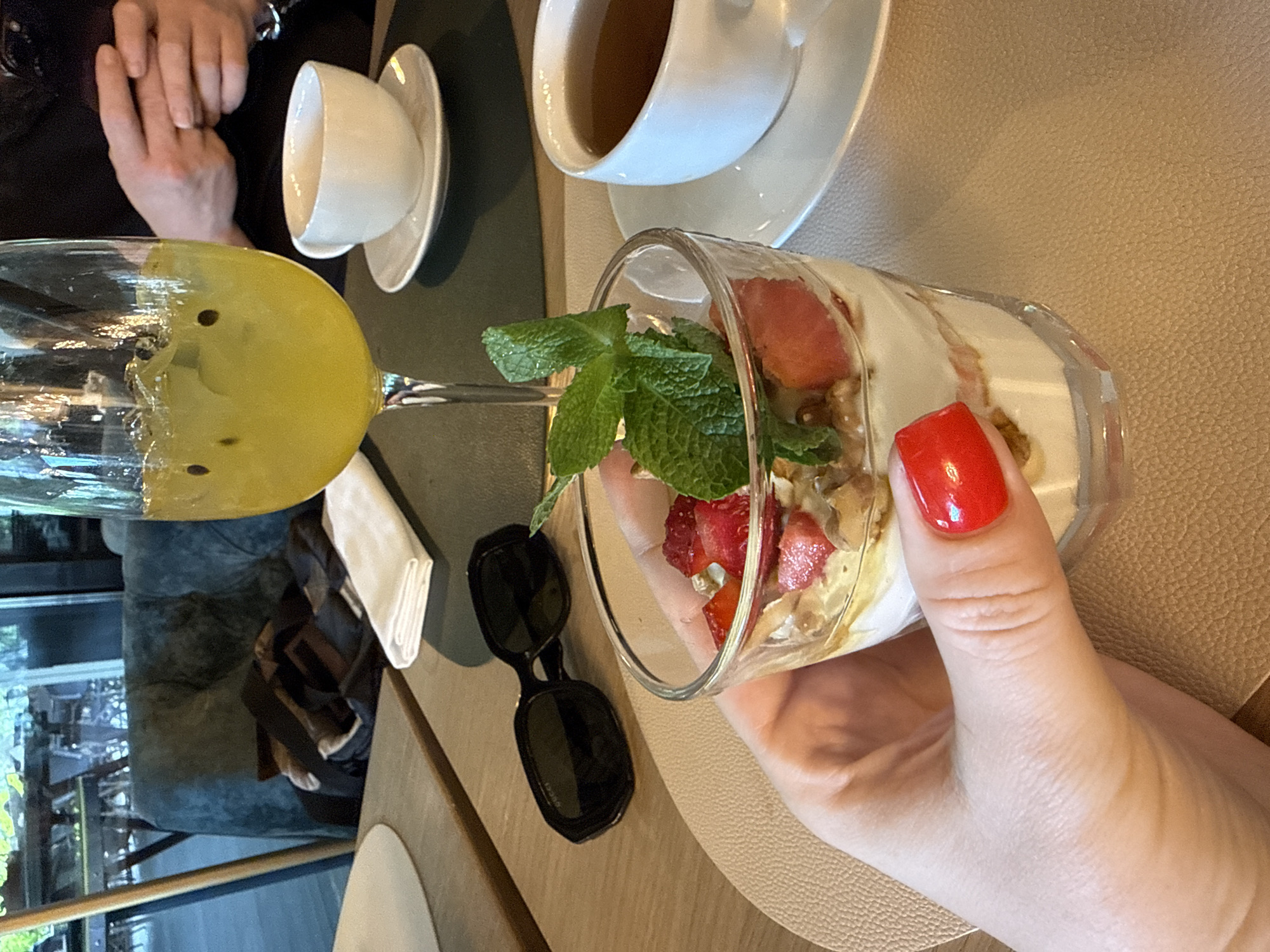 This media file captures a close-up, high-angle shot of a casual dining experience in what appears to be a cafe or restaurant in Moscow, Russia. The scene is brightly lit, suggesting daytime, with natural light entering from a large window in the background that shows reflections of greenery and what might be urban structures.
In the foreground, a hand with bright red nail polish holds a clear glass filled with a layered dessert. This dessert consists of white cream or yogurt, diced red strawberries, and what looks like granola or nuts, garnished with fresh mint leaves on top. To the left, a stemmed glass, possibly a wine glass, contains a yellowish drink with dark specks, perhaps a passion fruit-based beverage.
On the table surface, which is a textured light beige, are several items. A pair of black sunglasses, with the word "Gucci" faintly visible on the temple, rests near the dessert. A neatly folded white napkin is positioned further to the left. In the mid-ground, a white teacup filled with a dark liquid, likely tea, sits on its saucer. Another inverted white teacup on a saucer is visible in the upper left. Behind these, another person's hands are clasped together on a dark surface, possibly the table or their lap, indicating a companion at the meal. The background features plush dark blue or grey seating and a dark table, adding to the cafe ambiance. The overall atmosphere conveys a relaxed moment of indulgence.