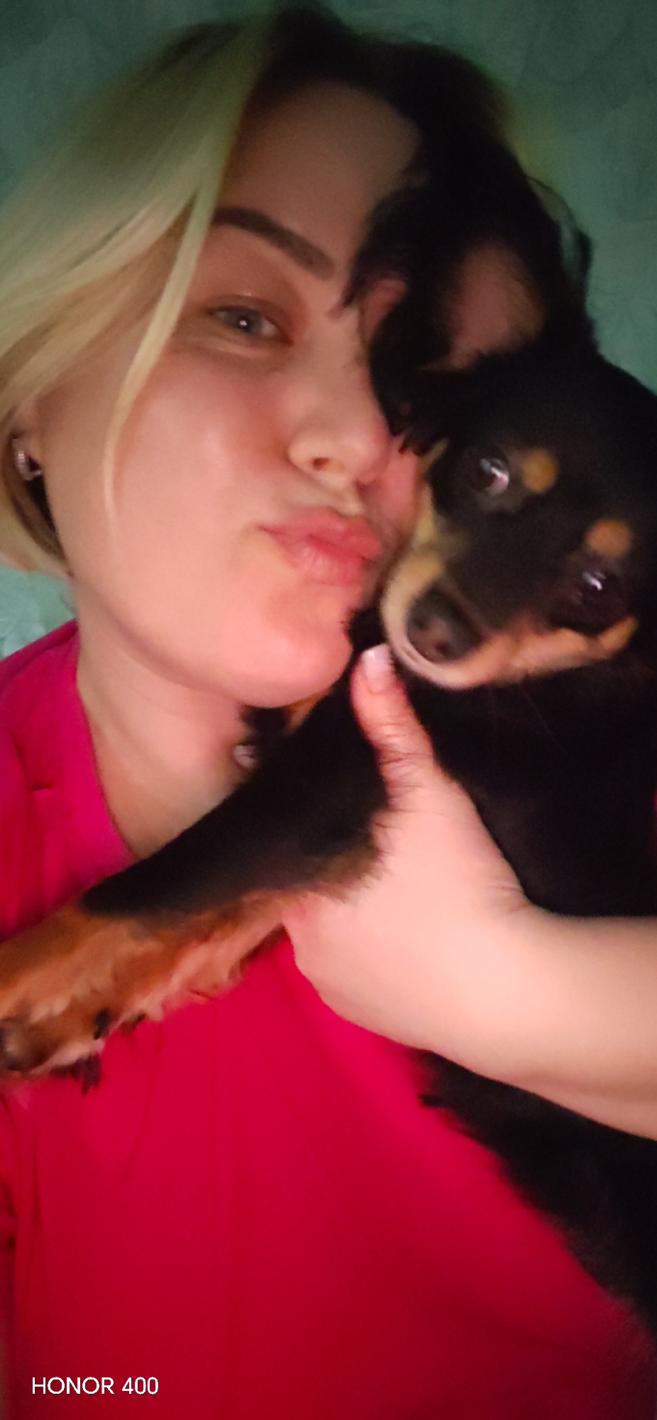 A woman with blonde hair and a red shirt holds a black and brown puppy. The woman is pursing her lips. The location may be Noviy, Russia. The photo appears to have been taken indoors and the words "HONOR 400" are visible at the bottom of the image.