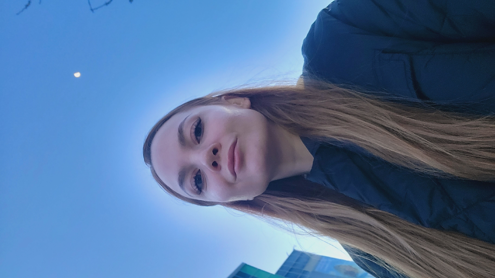 The image features a young woman captured in a selfie-style shot, with a low-angle perspective looking upwards towards a bright sky. The original image appears rotated, and when viewed correctly, the woman is positioned on the left side of the frame, with her head slightly tilted and her gaze directed towards the viewer.
She has long, light brown hair that flows around her face and over her shoulders. She is wearing a dark, possibly padded, jacket. Her facial features are clear, with well-defined eyebrows, noticeable eyelashes, and a subtle, closed-mouth smile that suggests a calm and pleasant demeanor.
The majority of the background is a clear, vibrant blue sky, indicative of fair weather. A distinct crescent moon is visible in the upper portion of the sky, suggesting the photo was taken during twilight hours, either early morning or late afternoon, when the moon can be seen against a still-bright sky. In the lower right corner, the edges of a modern building with reflective blue and possibly green-tinted windows are visible, hinting at an urban or developed setting, which aligns with the provided context of Chaikovsky, Russia. There are also some very faint, indistinct dark marks in the top left corner of the sky. The overall scene conveys a serene outdoor moment.