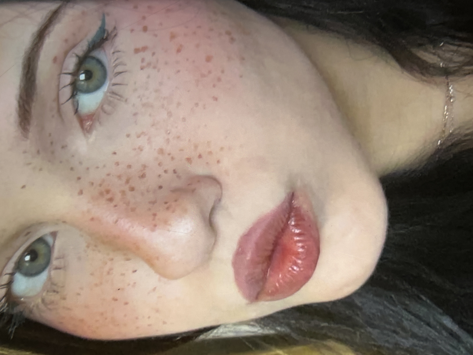 The media file is a close-up, upside-down shot of a person's face, focusing on their eyes, nose, and lips. The person has fair skin with prominent freckles scattered across their cheeks and nose. Their eyes are a striking light green, with long eyelashes and subtle blue eyeliner. A delicate silver necklace is visible on their neck. The lighting suggests an indoor setting, possibly a personal space, as there are no clear indications of the time of day or weather. The person's expression is difficult to ascertain due to the extreme close-up and unusual orientation, but the full lips, accentuated with gloss, could suggest a pose. No text is visible in the image. The context of Cupra Marittima, Italy, provides a geographical location but no direct visual cues within the image itself.