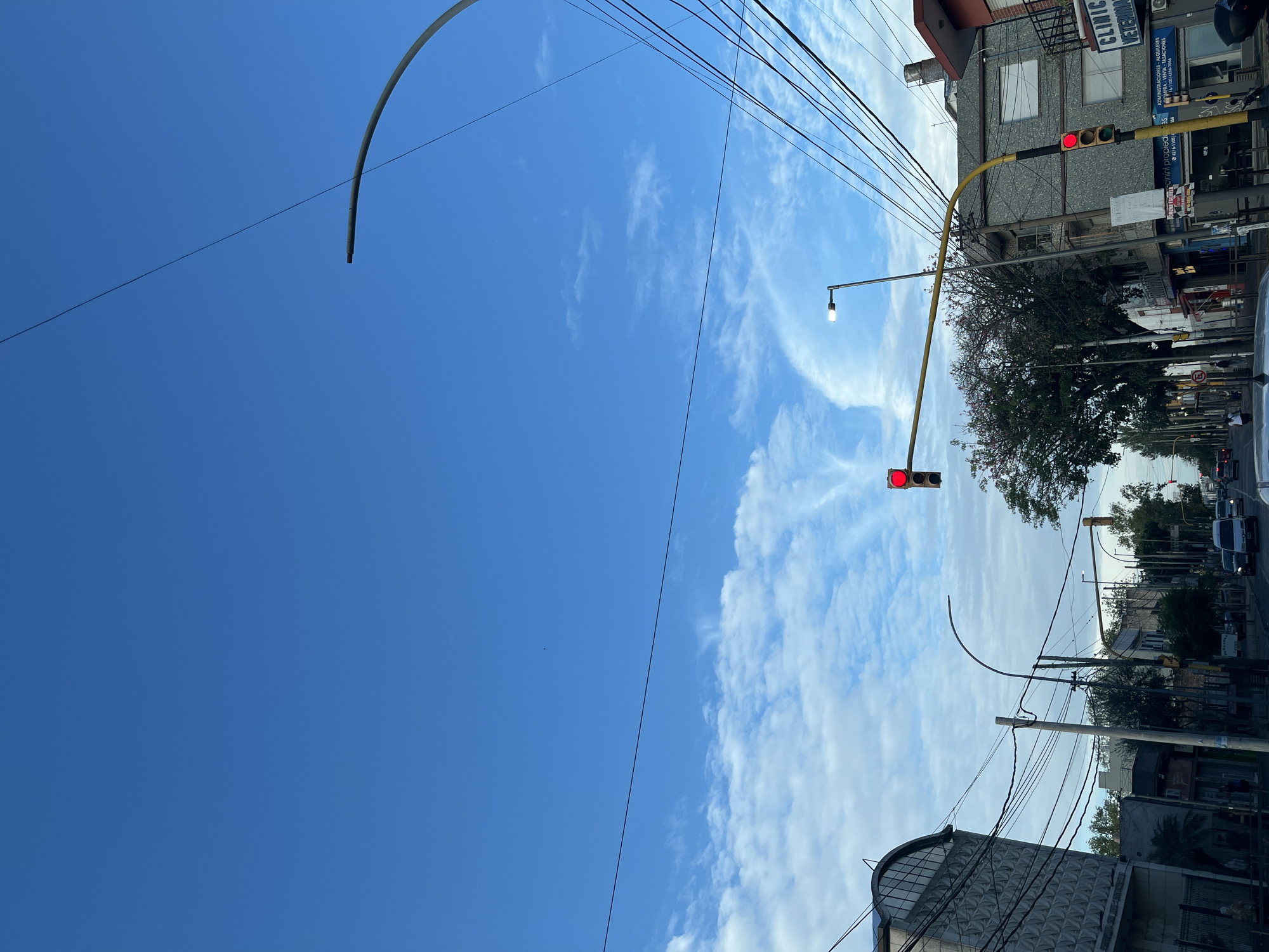 The image shows an upward-angled street view in Berazategui, Argentina, characterized by a clear blue sky interspersed with scattered white clouds. The scene is dominated by urban infrastructure, including overhead electrical wires and yellow traffic light supports. Two traffic lights are visible, both displaying red lights. A street lamp with a single bulb is also present. Buildings are visible along the street, some with visible signage, including a veterinary clinic. Trees are present, lining the street. Cars are visible on the street. The overall perspective suggests a residential or commercial area within the city.