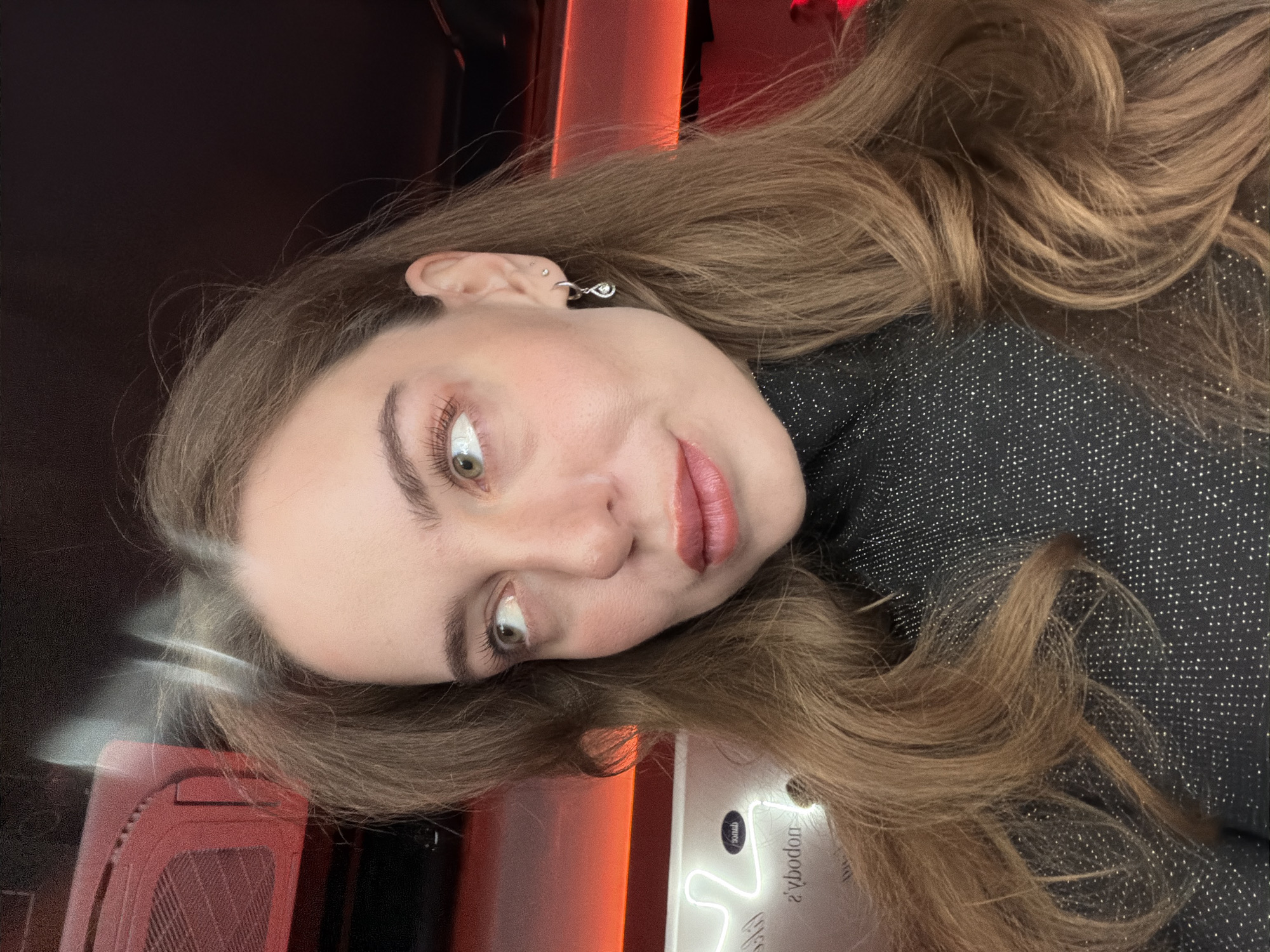 A female subject with long, wavy light brown hair and green eyes is depicted in a close-up, chest-up portrait. She is looking upwards and to the right of the frame, with her lips slightly parted and tinted peach/coral. She wears a dark, glittered turtleneck top and a silver teardrop-shaped dangle earring in her left ear lobe, along with a small stud in her upper cartilage. The background is dimly lit with red ambient strip lighting. A white neon sign displaying the mirrored text "žbodon" (reading "nobody's" when un-mirrored) is visible to the left of her head. A red rectangular object, possibly a ventilation unit, is partially obscured in the upper left. This indoor scene is identified as taking place at Nobody's Dance Bar, located at Ulitsa Pokrovka, 28с1, Moscow, Russia.