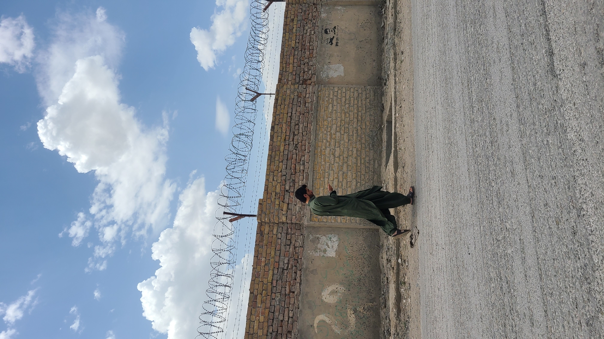 This image, taken in Quetta City Tehsil, Pakistan, captures an outdoor scene under a bright, sunny sky with scattered white cumulus clouds. The central figure is a man, viewed in profile, standing on an unpaved, dusty road composed of gravel and dirt. He is dressed in a dark green shalwar kameez and dark sandals, and has a beard and dark hair. His posture, with hands raised and gaze directed upwards, suggests an act of prayer or deep contemplation.
To the man's left, a long, weathered wall dominates the midground. The upper section consists of reddish-brown bricks, topped with a dense line of coiled barbed wire fencing that runs horizontally across the frame. Below the brickwork, the wall transitions into a lower concrete section, which appears aged and bears various markings. Notably, a dark stencil on the concrete features a stylized image resembling a skull or face, accompanied by some barely legible dark text, which seems to include characters like "WI-34". Further down on the concrete, faint white markings, possibly symbols or numbers, are visible. The overall environment conveys a sense of a rugged, possibly semi-urban or rural setting.