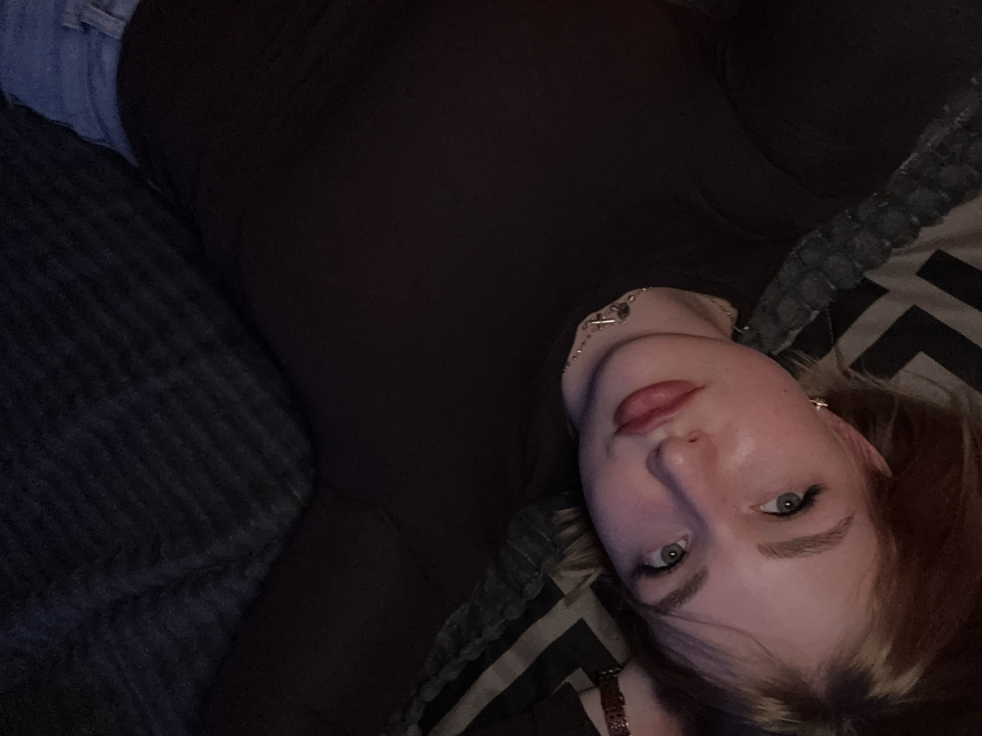 A young Caucasian female is lying on a dark-colored blanket with a striped pattern. She is looking upwards towards the camera, with her head tilted to the side. Her hair is light brown and falls around her face. She is wearing a dark shirt and a necklace with a cross pendant. Her makeup includes eyeliner and lipstick. The lighting in the image is dim, suggesting an indoor setting. The location in the city of Khimki, Russia, is not discernible from the provided image.