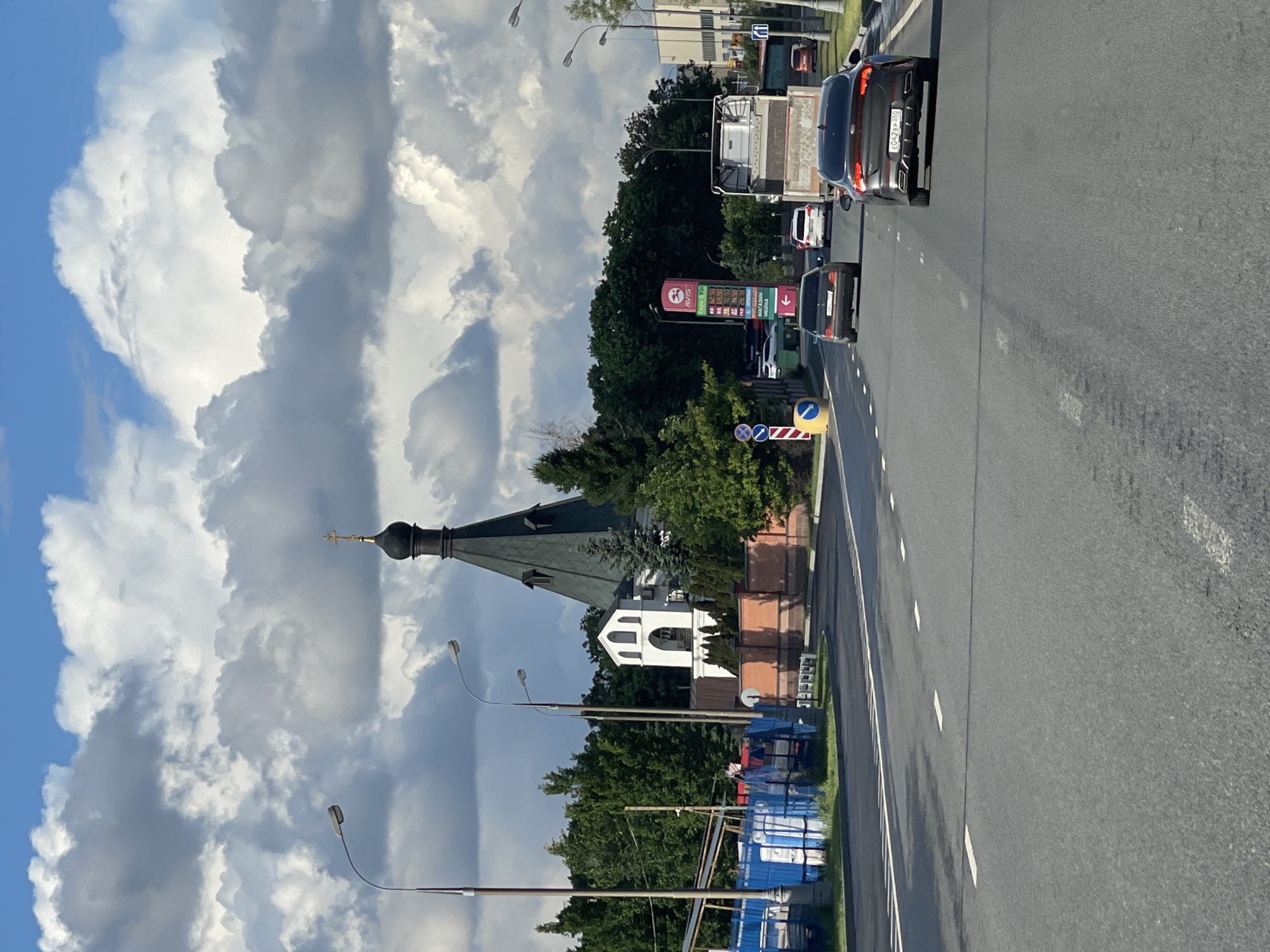 The image shows a street scene in Bittsa, Russia. On the left side of the road is an Orthodox church with a black dome and gold cross, partially obscured by trees. A large, cloudy sky dominates the background. The right side of the image is dominated by a gray asphalt road with white lane markings. There are several vehicles on the road, including cars and a truck. A gas station sign, labeled "AVIS", is visible amidst green foliage. The scene suggests a mix of urban and religious elements in the Bittsa landscape. Construction materials are present near the church.