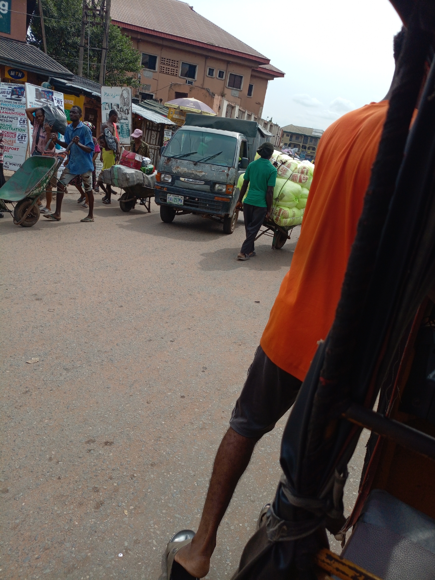 The image captures a bustling street scene, likely in a market or commercial district of Onitsha, Nigeria, taken from the perspective of a person in a vehicle, possibly a tricycle, evidenced by the visible leg, foot, and parts of a seat and frame on the right.
The street is a hive of activity, with several people engaged in transporting goods. Multiple individuals are seen pushing or standing near wheelbarrows, some empty and others heavily loaded. On the left, a man in a blue shirt pushes a green wheelbarrow. Further along, another wheelbarrow is piled high with bags and sacks. A small, dark-colored truck or van, bearing the words "TWIN CAM" and a Nigerian license plate "ENU-253MK", is visible in the middle of the road. To the right, a man in a green shirt pushes a wheelbarrow overflowing with numerous large, light-colored sacks, likely containing agricultural products.
Buildings line the street in the background. On the left, a "BLESSED COMPUTER SERVICES OFFICE" displays banners advertising services such as "PHOTOCOPY," "TYPING," "MAINTENANCE," "GRAPHICS CBT," "DESIGN," "ONLINE REGISTRATION," and "RESULT CBT." An "MTN" sign is also visible above the computer office. Another banner displays "Pargon," and an orange umbrella with the partial text "GLOBAL CONCE" is seen in the distance. The scene unfolds during daylight hours under a bright, possibly overcast sky, conveying the busy nature of commerce and daily life in the area.