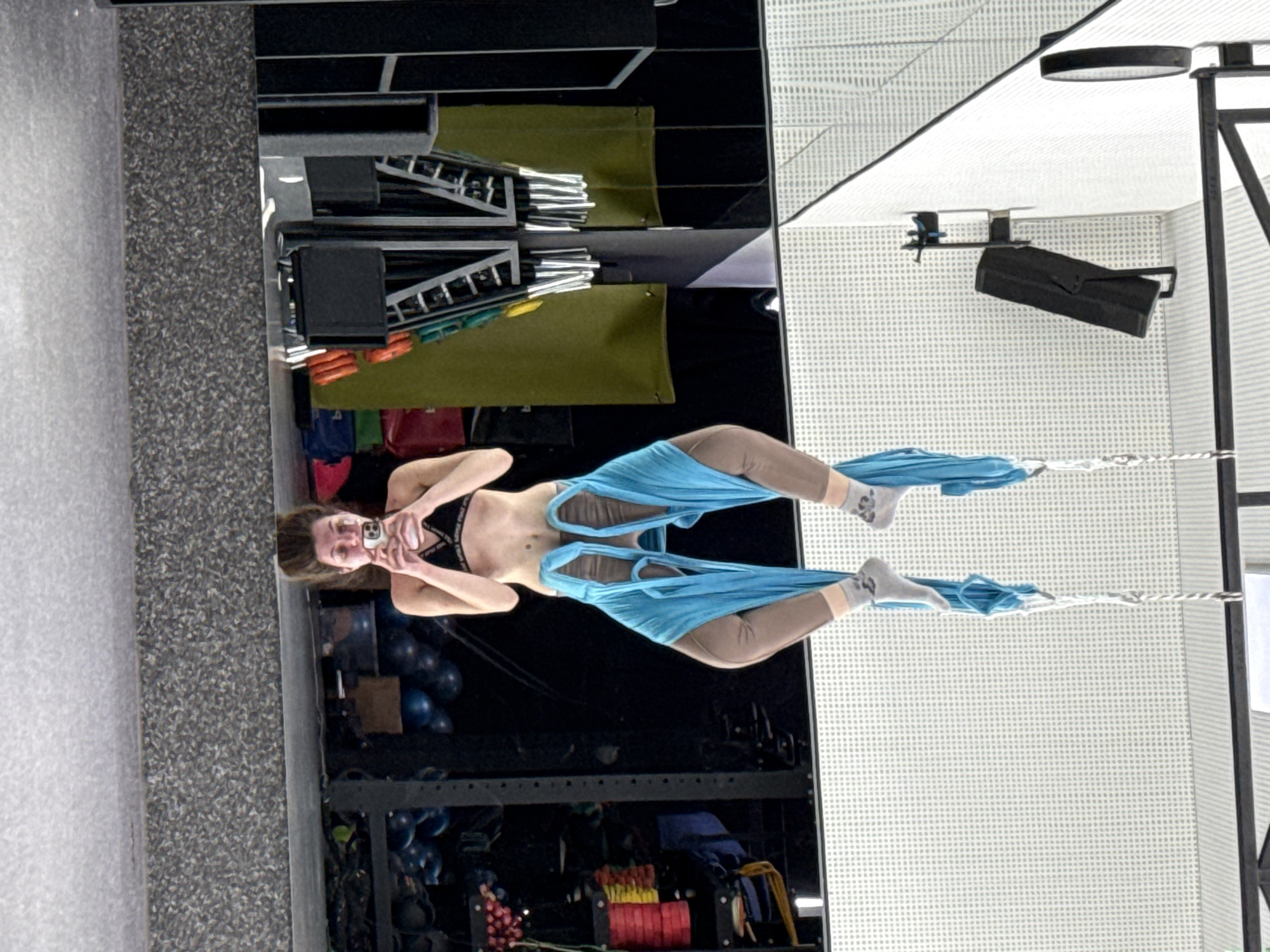Here is a detailed description of the image:
**Scene:** An indoor gym or fitness studio.
**Subject:** A female individual is suspended in the air using aerial silks or hammocks. She is wearing athletic attire, including a sports bra and leggings. She is holding a phone and taking a selfie.
**Objects:**
* Aerial silks/hammocks in a light blue color.
* A phone used for taking a selfie.
* Fitness equipment such as weights, racks, and exercise balls are visible in the background.
* Lighting fixtures and ceiling structures are present.
**Actions:**
* The woman is performing aerial yoga or a similar fitness activity.
* She is holding a phone and taking a selfie.
**Interactions:**
* The woman is interacting with the aerial silks/hammocks to perform the exercise.
* She is interacting with her phone to take a photo.
**Environment:** The background features typical gym equipment and the studio seems well-lit.
**Location:** The city and region are not discernible.