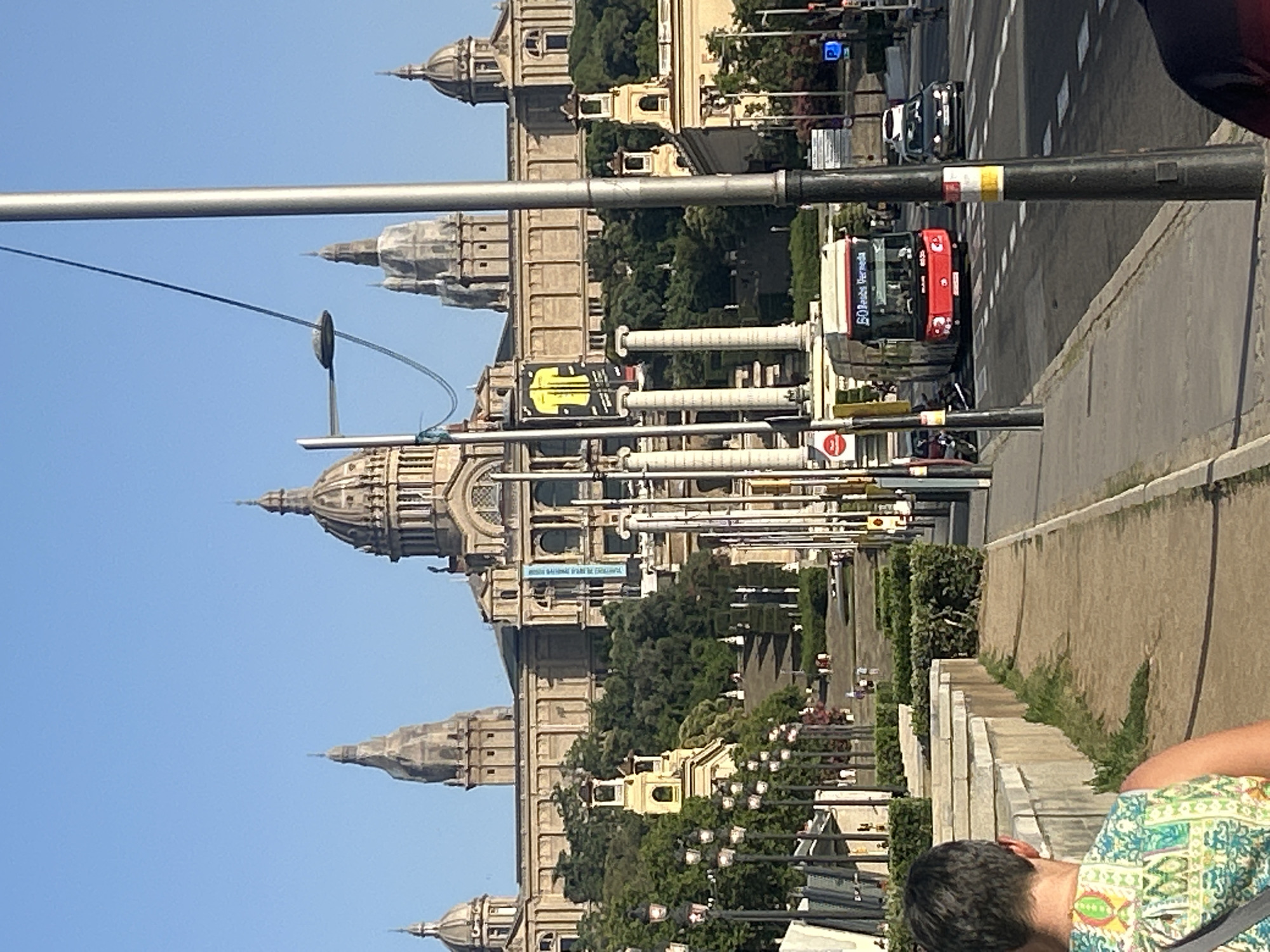 The image shows a view of the Palau Nacional in Barcelona, Spain, with its grand facade and domes dominating the skyline. In the foreground, a street with traffic is visible, including a red bus labeled "50 Barceloneta Verneda" and a black car. A person's back is in the lower left corner, wearing a colorful patterned top. The Palau Nacional is set against a clear blue sky, with trees and manicured hedges lining the approach to the building. The architecture is neoclassical, with grand columns and ornate details. Streetlights line the sidewalk on the right side of the image. The scene appears to be captured during the daytime.