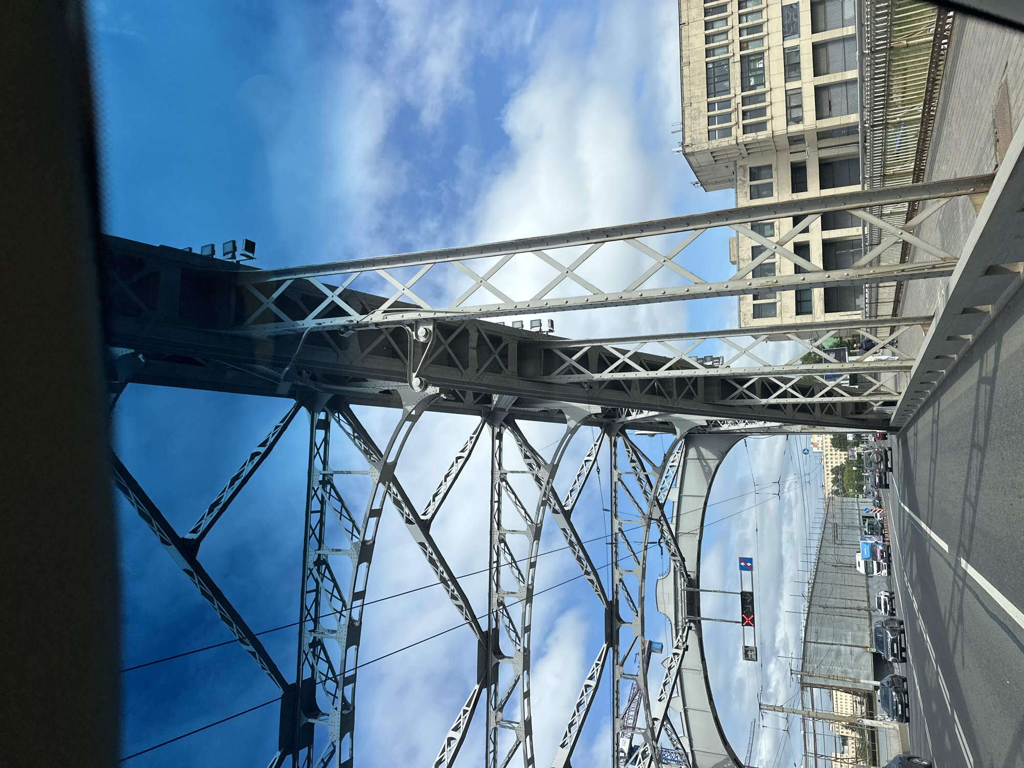 The image shows a view from a vehicle driving across the Peter the Great Bridge in Saint Petersburg, Russia, on a partly cloudy day. The bridge's distinctive grey metal truss structure dominates the foreground and mid-ground, with its complex network of beams and supports visible against the bright blue sky and scattered white clouds. In the distance, several multi-story buildings with a utilitarian architectural style are visible. The bridge carries vehicular traffic, with several cars and trucks driving on the roadway. Traffic signals and overhead wires are also present. The perspective suggests the image was taken from the driver's or passenger's seat of a car.
