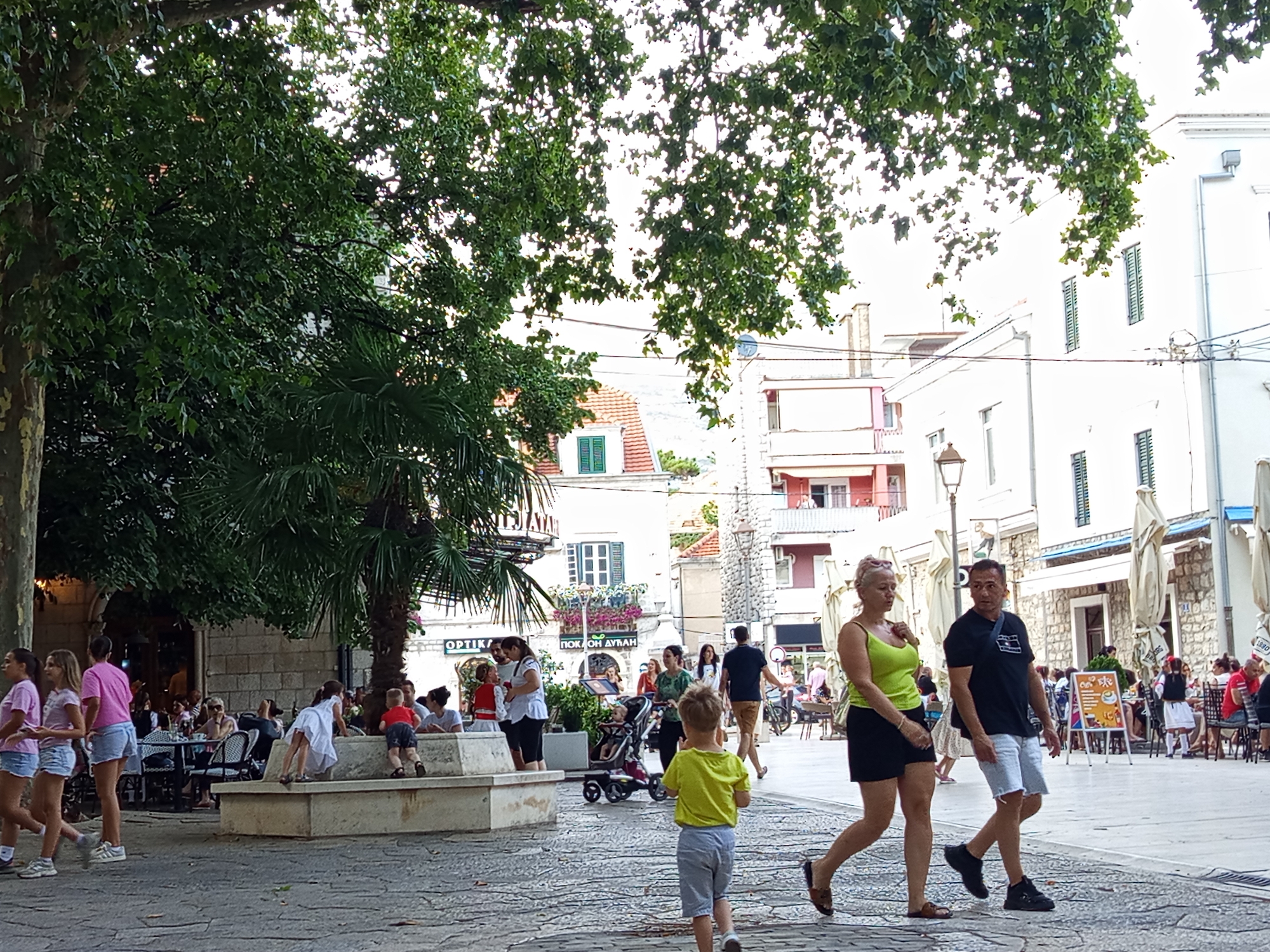The image depicts a lively public square in Trebinje, Bosnia and Herzegovina. The scene is bustling with activity, featuring a mix of adults and children engaged in various pursuits. In the foreground, three young girls in matching pink shirts and denim shorts walk together from left to right, with the leftmost girl looking towards the camera. To their right, a group of people are seated at tables outside a restaurant, some engaged in conversation, others appearing to dine. A young boy in a yellow shirt and grey shorts is walking away from the camera towards a concrete structure. Behind him, a young girl in a white dress is interacting with a child on the concrete structure. Further back, a couple walks from right to left, the woman in a green top and black skirt, the man in a black t-shirt and white shorts. The square is paved with cobblestones, and large trees with dense foliage provide shade. Buildings with white facades and red-tiled roofs line the square, some featuring balconies with potted plants. There are outdoor seating areas, umbrellas, and streetlights visible. Signs in what appears to be Cyrillic script are present on some buildings, indicating local businesses. The overall atmosphere suggests a warm, pleasant day in a well-frequented urban public space.