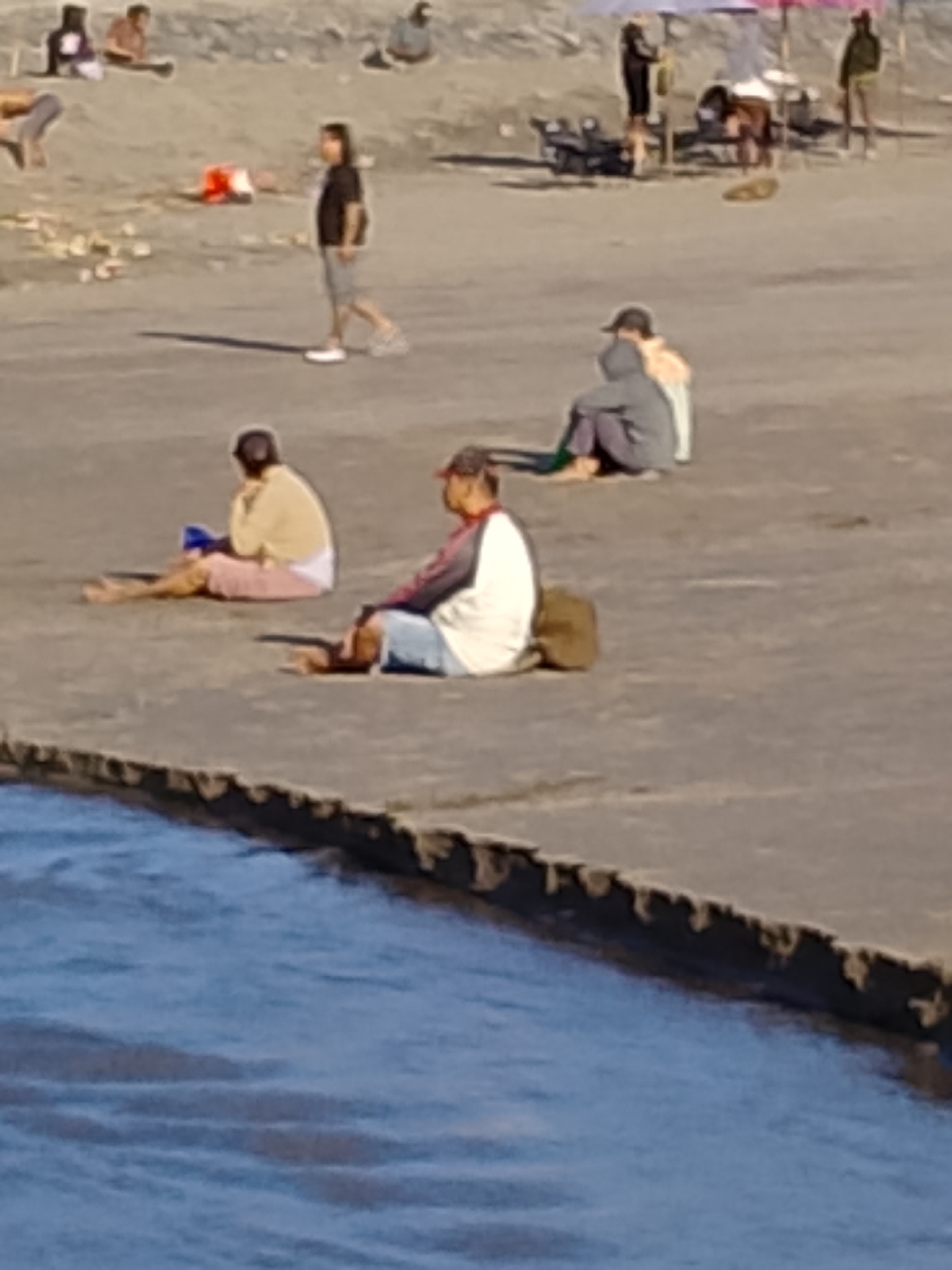 In the city of Kediri, Indonesia, a sandy riverbank is occupied by several individuals. In the foreground, a man in a red-capped baseball cap and a white and red jacket sits facing left, with a brown bag beside him. To his left, a woman in a yellow top and pink pants is also seated. Further up the bank, a person in a dark shirt and grey shorts walks away from the viewer. In the background, a person wearing a grey hoodie sits facing away from the viewer. Other figures are visible further back, some near a beach umbrella and a motorcycle. The river flows along the bottom edge of the image.