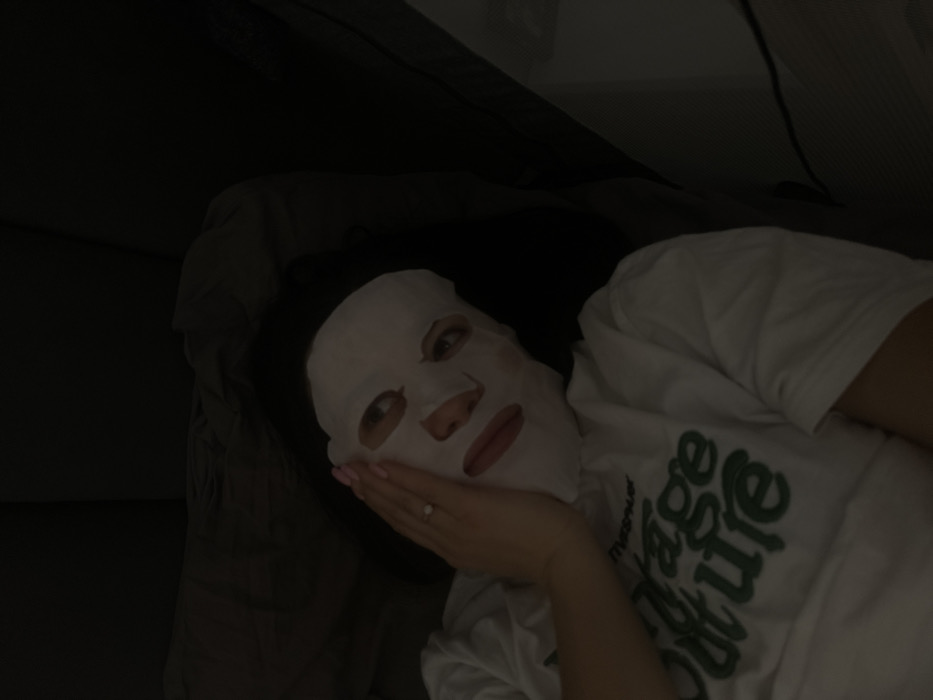 An individual is depicted lying on their back in a dimly lit indoor environment in Zhukovka, Russia. The person, appearing to be an adult female with dark hair, has a white sheet mask applied to their face, covering the forehead, cheeks, and chin, with cutouts exposing the eyes, nose, and mouth. The eyes are open and directed slightly towards the camera, while the mouth reveals red-toned lips. The individual's left hand, with light pink painted fingernails and a ring featuring a clear stone on the ring finger, is placed flat against their left cheek. They are wearing a white short-sleeved t-shirt with partial green lettering visible on the chest, appearing to read "Vintage Couture". The background consists of dark, solid-colored fabric, indicative of a bed or sofa. No interactions are visible.