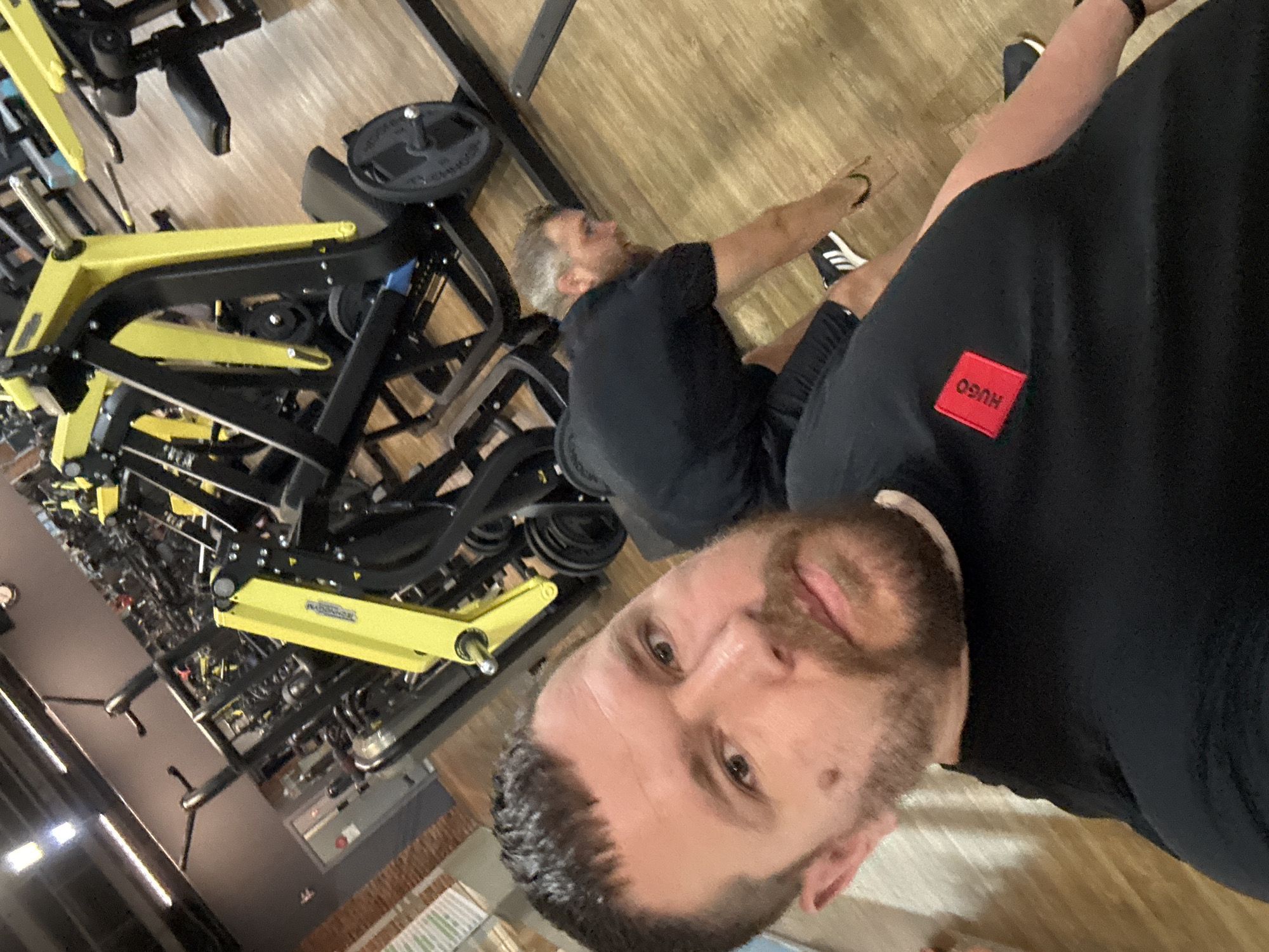 The image is a selfie taken in what appears to be a gym. The location is not confirmed as Chaikovsky, Russia.
Foreground: A man with short dark hair and a beard occupies the lower right portion of the image. He is wearing a black shirt with a red "HUGO" label.
Midground: Behind the man, another man is sitting on the floor. He is wearing black shorts and a black shirt. He is partially visible and appears to be gesturing with his right hand.
Background: Gym equipment dominates the background. There are yellow and black weight machines and various other fitness devices. The floor is made of wood. The lighting is bright and appears to be artificial.