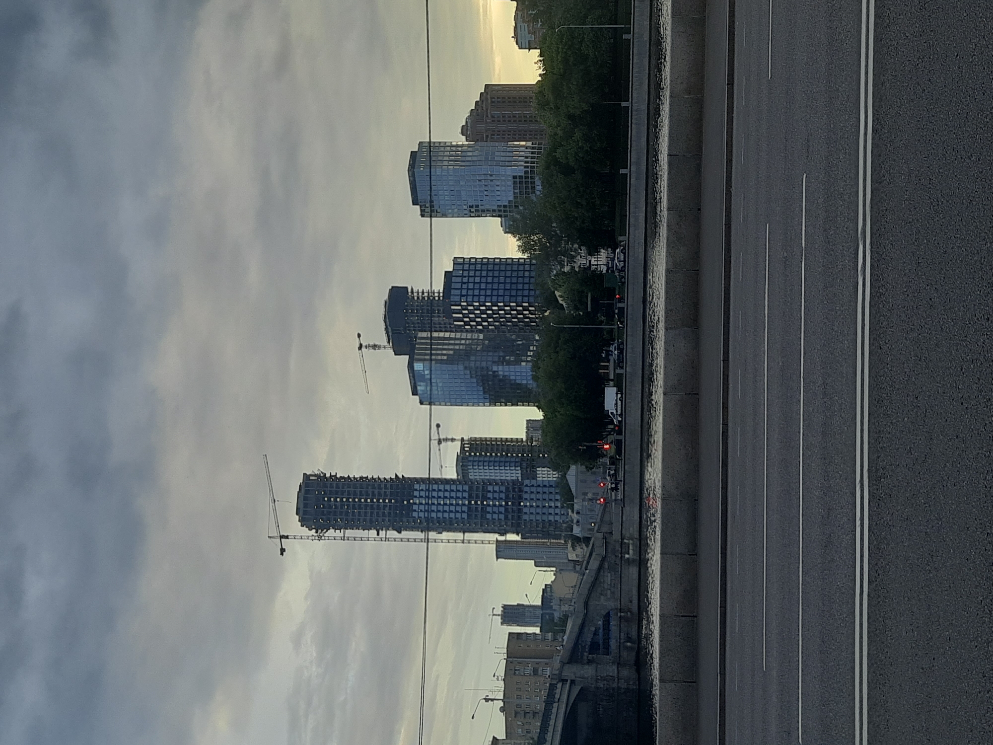 This image, oriented vertically, captures a cityscape with a prominent road in the foreground. The road, paved with asphalt, is marked with white lines, indicating traffic lanes. To the left of the road, a concrete barrier separates it from a view of buildings under construction and completed high-rises.
Several multi-story buildings are visible, some still in their skeletal form with visible construction elements like cranes, while others appear complete with glass facades reflecting the sky. A line of green trees stands between the construction and the road, suggesting a park or urban greenery. The sky is overcast with clouds, hinting at a subdued atmosphere. The light suggests it is either early morning or late evening.
The scene appears to be an urban environment, likely a city, given the presence of tall buildings and a major road. The construction activity indicates ongoing development. There are no visible people or specific objects that stand out, other than the architectural elements and the road infrastructure. The overall mood is neutral, focusing on the urban landscape and its development. The context provided indicates the location as Moscow, Russia.