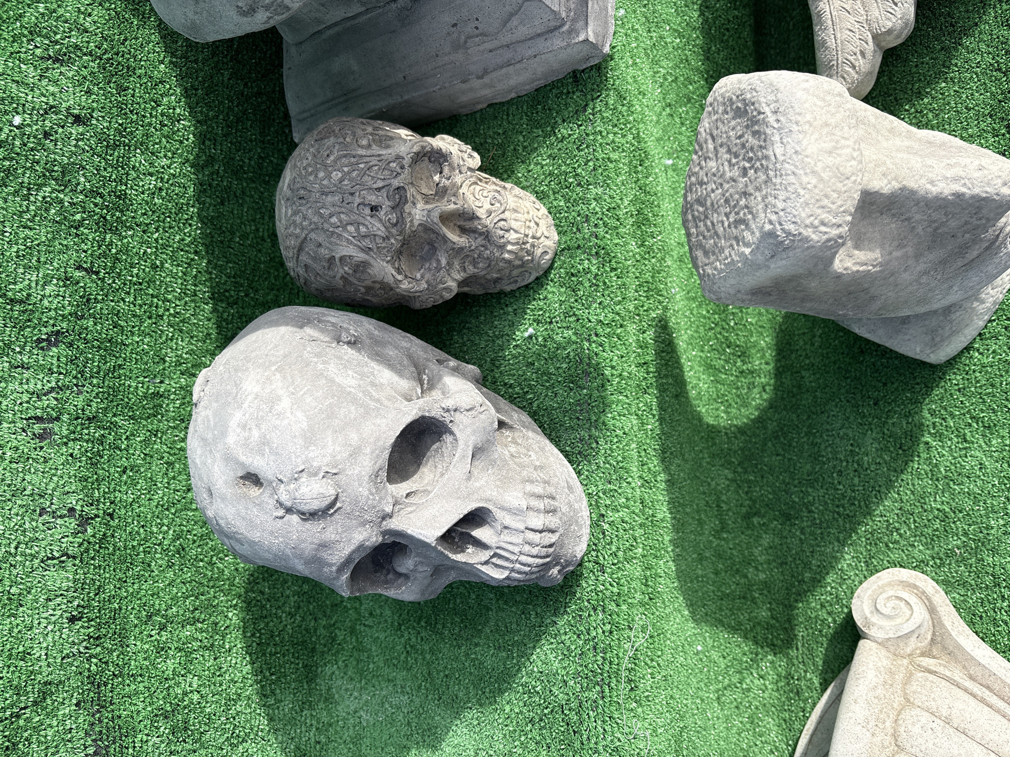 The image depicts a collection of stone skull sculptures, along with other architectural fragments, lying on a vibrant green surface resembling artificial grass. The scene is brightly lit, casting distinct shadows from the objects onto the green surface. The skulls vary in design; one has intricate patterns carved into its surface, while the others are more plain. The other objects appear to be architectural elements, such as pieces of columns or capitals. The image provides no information about whether this is in Chirk or anywhere else.