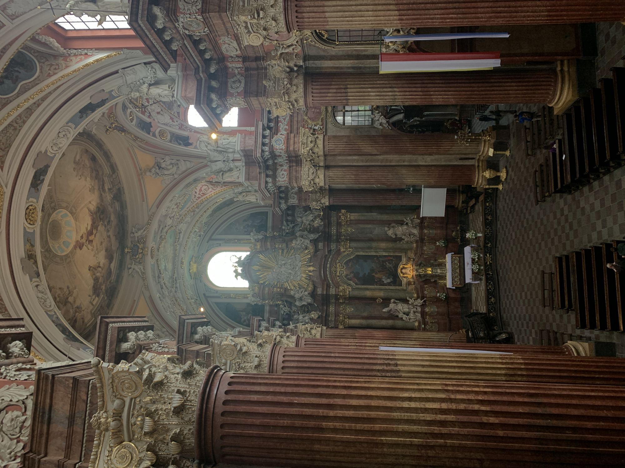 The image shows the interior of St. Stanislaus Church in Poznan, Poland. The church is ornately decorated in a Baroque style, with gilded accents, intricate carvings, and colorful frescoes on the ceiling. Large, fluted columns with elaborate capitals line the nave. The altar area features a large painting and numerous sculptures. Benches for congregants are arranged in rows on the checkered floor. A few people are visible in the church, including some seated on benches and others standing near the altar. The lighting is dim, with natural light entering from arched windows.