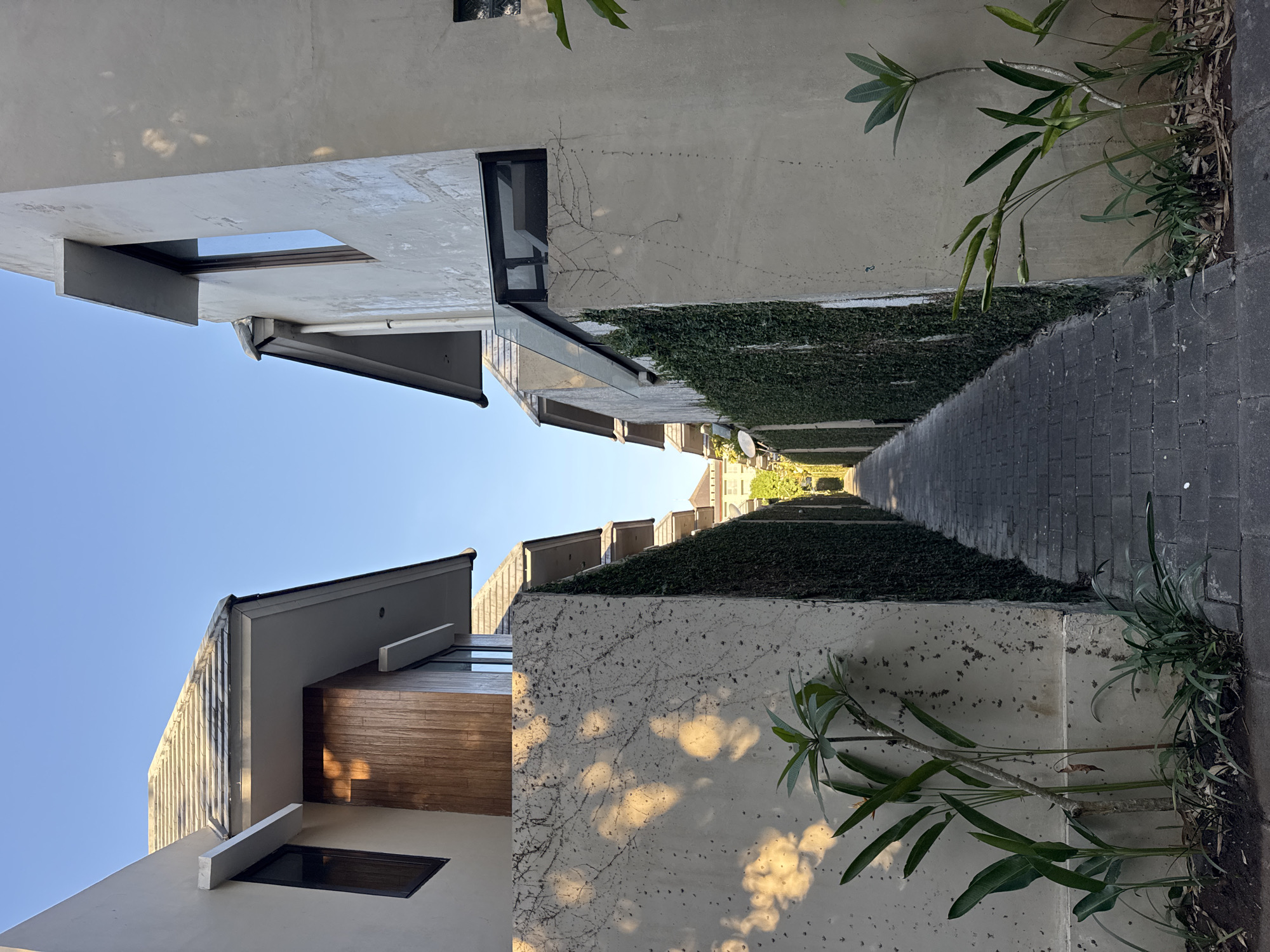 The image shows a narrow paved pathway between two buildings. The pathway is made of dark grey rectangular pavers. On either side of the pathway are tall, light-colored walls covered in dense green ivy. The walls are approximately 2-3 meters high. Above the walls, the upper portions of the buildings are visible. The building on the left has a tan-colored exterior and a dark wooden accent wall. It features a light-colored cantilevered roof overhang and a dark-framed window. The building on the right also has a light-colored exterior, a dark-framed window, and a modern design with a glass awning. The sky is clear and blue. In the foreground on the left, there are several green plants growing near the wall, and on the right, there are also some plants. Sunlight casts dappled shadows on the walls and pathway. The scene appears to be a quiet, residential or resort-like area in Kuta Selatan, Indonesia, likely in a private villa complex given the architecture and landscaping. No people are present in the image.