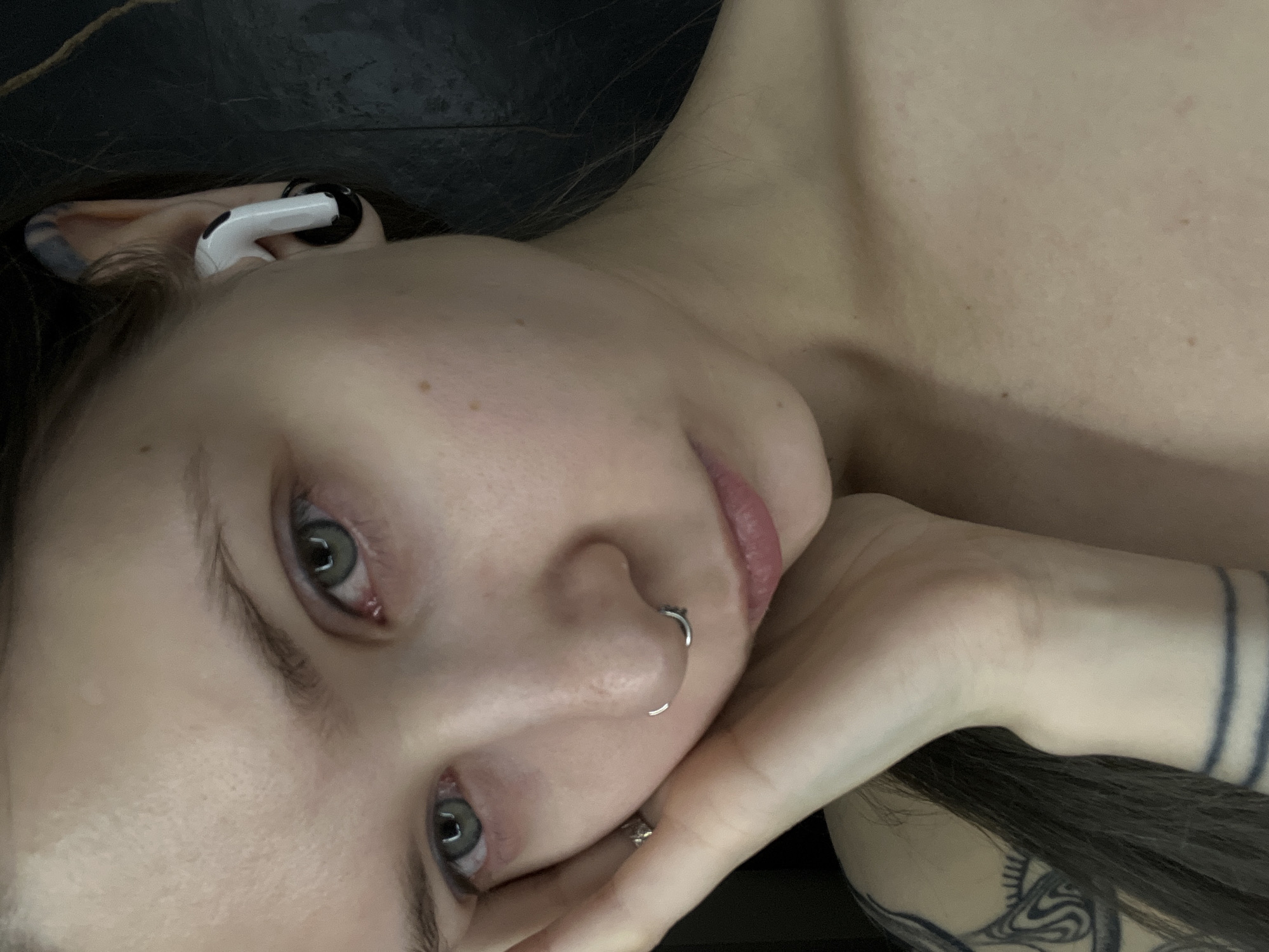 A close-up, chest-up portrait depicts a young adult female with fair skin, light green eyes, and long dark hair. She gazes directly forward with a neutral expression. Her left hand rests against her left cheek and jawline, displaying a ring on her ring finger. Her nose features a silver circular barbell septum piercing and a small silver hoop in her left nostril. A white wireless earbud, with a black accessory attached to its stem, is visible in her right ear, which also shows a blue/purple tattoo or discoloration in the concha area. Tattoos include an intricate black line-art design on her upper left shoulder/back and three horizontal black bands on her left wrist/forearm. Her upper chest and shoulders are bare. The background is a dark, non-descript surface, indicating an indoor setting. This scene is located in the city of Khimki, Russia.