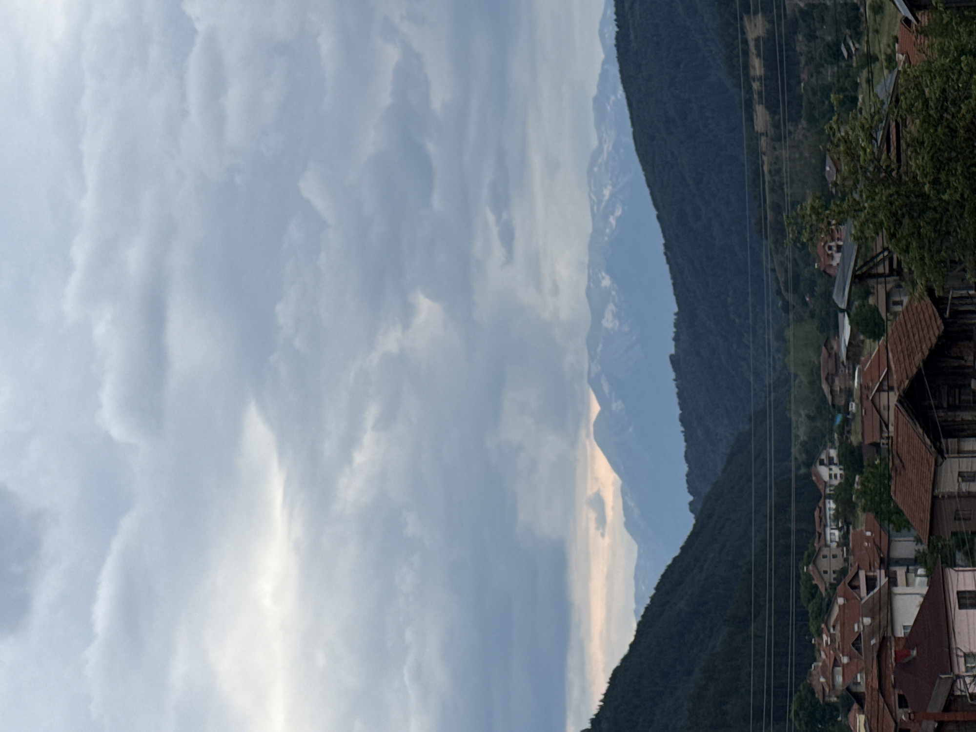 The image shows a village nestled in a valley with mountains in the background. The sky is overcast with thick clouds. The village appears to be comprised of houses with red tile roofs. Some houses are partially obscured by trees. In the foreground, several houses are visible, some with weathered facades. Power lines stretch across the scene, suspended between poles that are not fully visible. The mountains in the background are covered in dark green vegetation on their lower slopes and have snow-capped peaks. The overall impression is one of a rural, mountainous landscape under a cloudy sky.