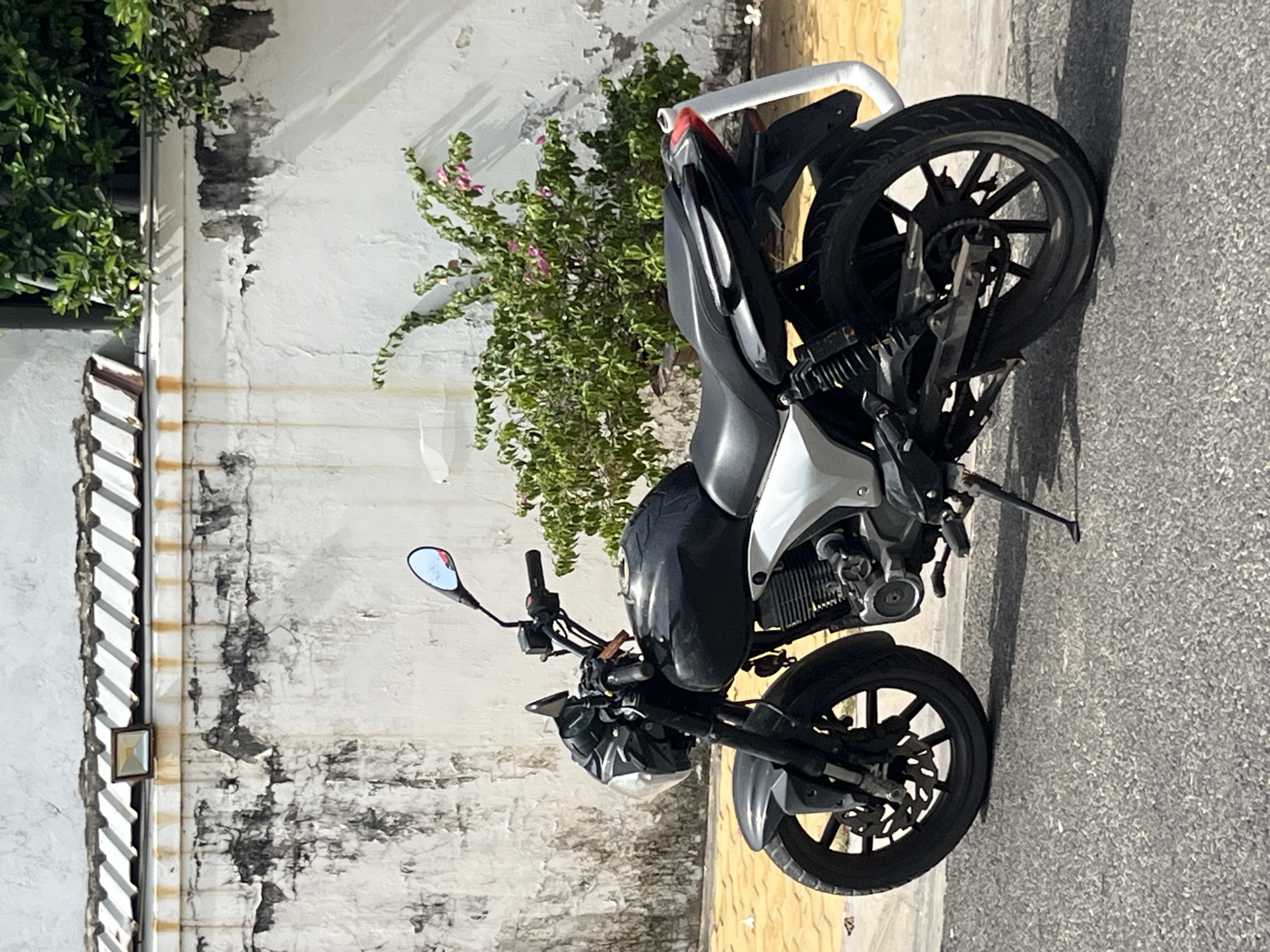 A black and silver motorcycle is parked on an asphalt road next to a yellow tiled sidewalk in Mũi Né, Vietnam. The motorcycle is facing left, with its kickstand down. Behind the motorcycle is a white stucco wall with peeling paint and rust stains. A corrugated metal roof runs along the top of the wall. To the right of the motorcycle, a dense green bush with pink flowers is growing. The sky is not visible in the frame.