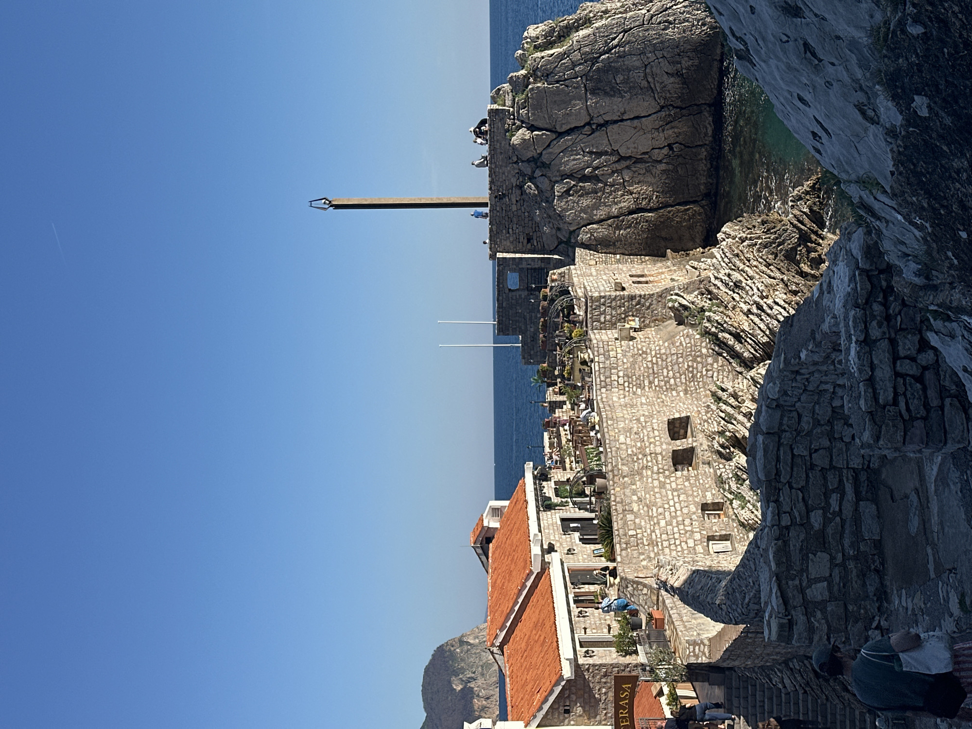 This image shows a view of the city of Petrovac centar, Montenegro, with a clear blue sky above. Visible elements include stone buildings with terracotta roofs, a stone monument on a rocky outcrop, a body of water that appears to be the Adriatic Sea, and stone formations. Some people are visible on the lower right edge of the photo. The image is taken at a slight angle.