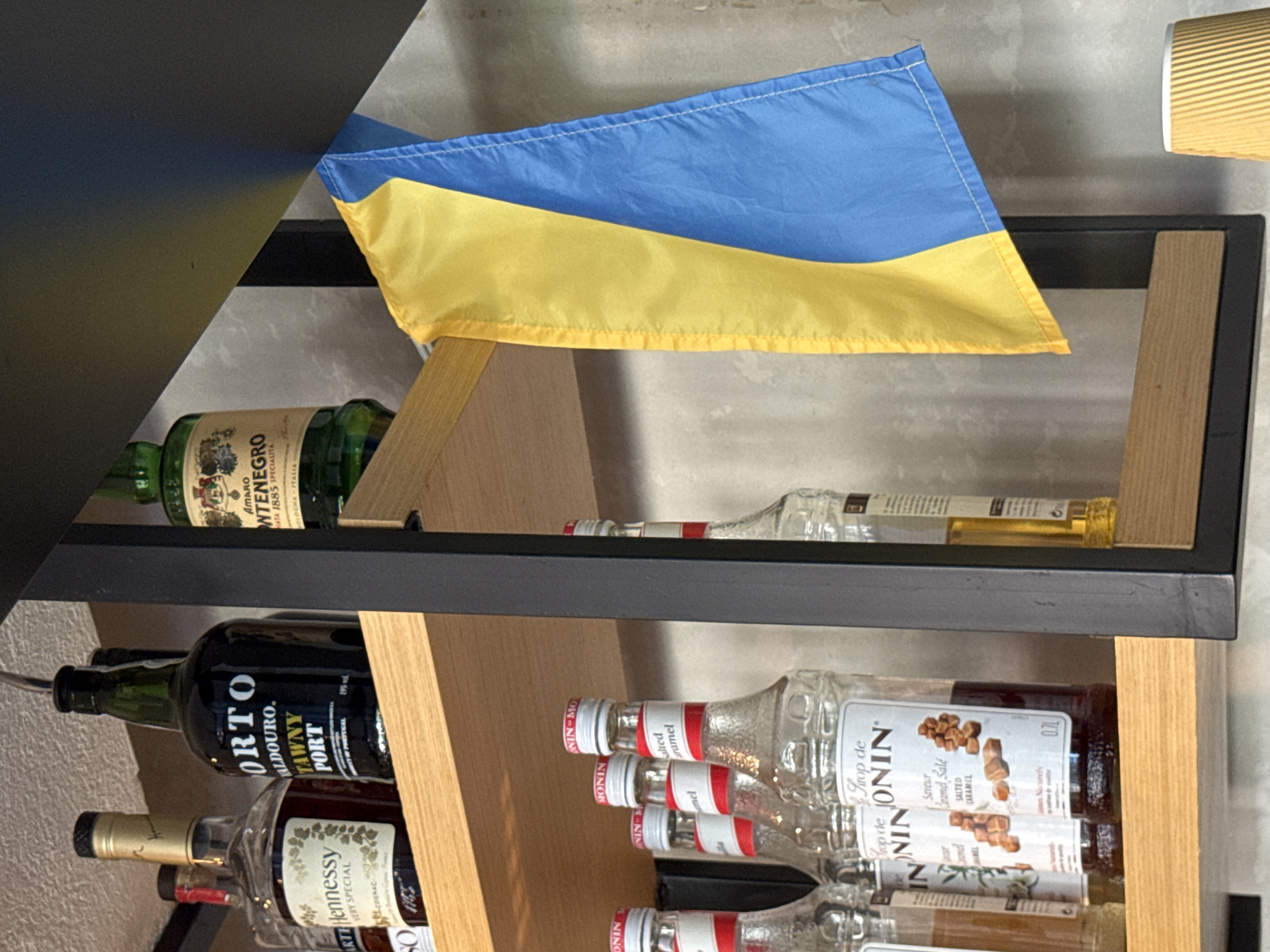 The image displays a shelf in what appears to be a bar or cafe. On the top shelf, there are several bottles of alcohol, including a bottle labeled "PORTO VALDOURO TAWNY PORT" and another with "AMARO MONTENEGRO". A Ukrainian flag is draped over the edge of the shelf in the middle section of the image. The lower shelf holds multiple bottles of flavored syrups, identifiable by the "MONIN" branding and labels such as "Salted Caramel". The background appears to be a textured wall. The scene is indoors, with artificial lighting. The location is identified as Boreti, Montenegro, though this context is not visually evident in the provided image.