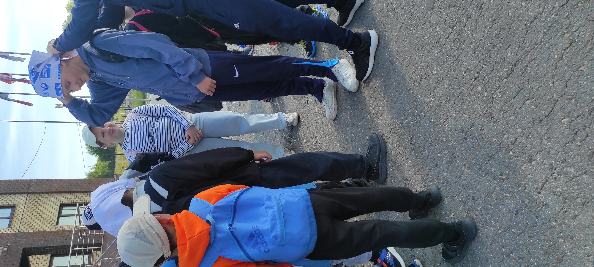 The image shows a group of people, likely children and teenagers, standing outdoors on an asphalt surface. Some of the individuals are wearing athletic clothing, including tracksuits and sneakers. One person is adjusting a white bandana on their head, which has blue text and logos on it. Another person in the foreground is wearing a bright blue backpack with the Adidas logo visible. The scene appears to be set in a public area, possibly near a building with brickwork and windows, and a fence. There are also flags or banners visible in the background. The lighting suggests it is daytime, with a clear sky visible. The overall mood seems casual, perhaps related to an outdoor activity or event. The visible text on the bandana appears to be in a Cyrillic script, with the words "ГАЗПРОМ" (Gazprom) and potentially "НЕФТЕХИМ" (Neftekhim) visible. The setting is identified as Noviy, Russia.