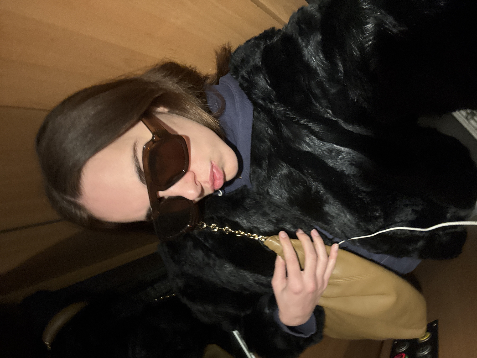 A woman with brown hair and brown sunglasses is wearing a dark blue hoodie and a black fur coat. She is holding a tan leather purse with a gold chain strap. The background appears to be wood paneling. The location cannot be determined to be Paris, France based on the image.
