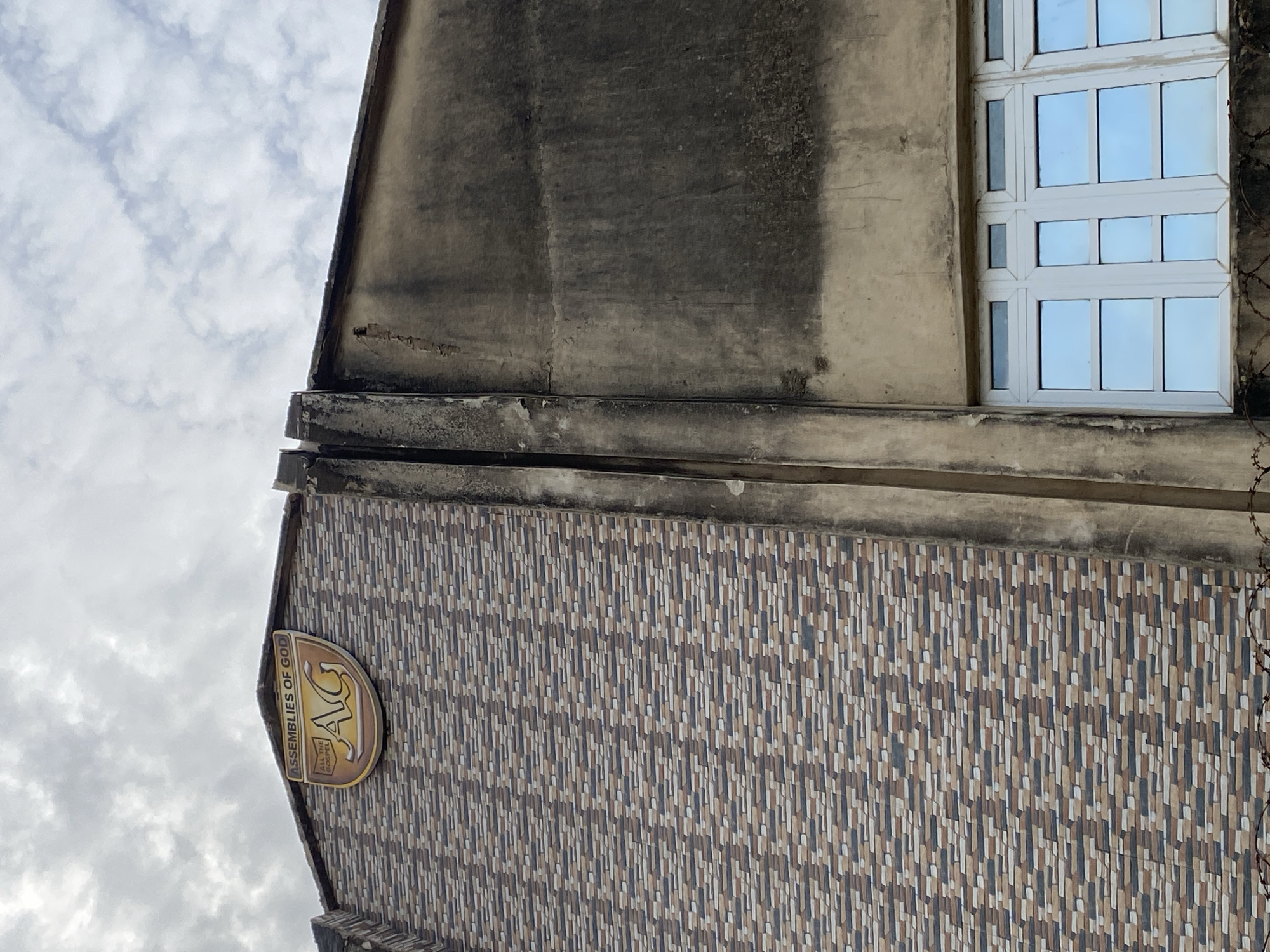The image shows the exterior of a building in Jos, Nigeria. The building has a tiled facade on the left side, with a logo that reads "ASSEMBLIES OF GOD" and "AG" with the slogan "ALL THE GOSPEL". The right side of the building is a concrete wall that appears weathered and stained. A white-framed window with multiple panes is visible at the bottom right. Barbed wire is present along the bottom edge of the image. The sky is cloudy.
