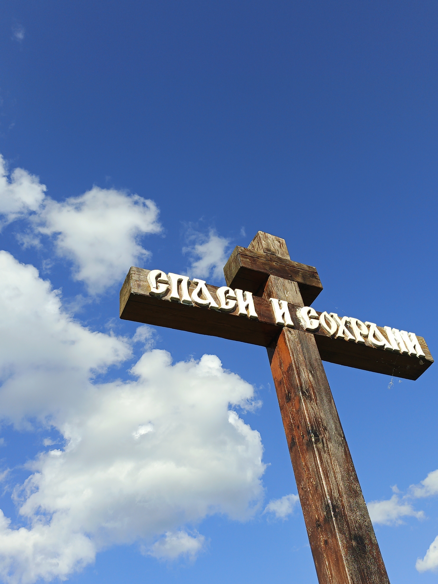 A close-up, low-angle shot shows a weathered wooden cross against a bright blue sky with scattered white clouds. The cross has white, raised lettering on its horizontal beam. The lettering appears to be in Cyrillic script and reads "СПАСИ И СОХРАНИ" which translates to "Save and Protect". The cross is positioned at an angle, with the main upright beam extending downwards out of frame to the right, and the horizontal beam extending to the left. The sky is clear and vibrant, with fluffy white cumulus clouds drifting across it. The overall impression is one of a religious symbol set against a vast and open sky, suggesting a location of spiritual significance. The image is from Maly Berdyaush, Russia.