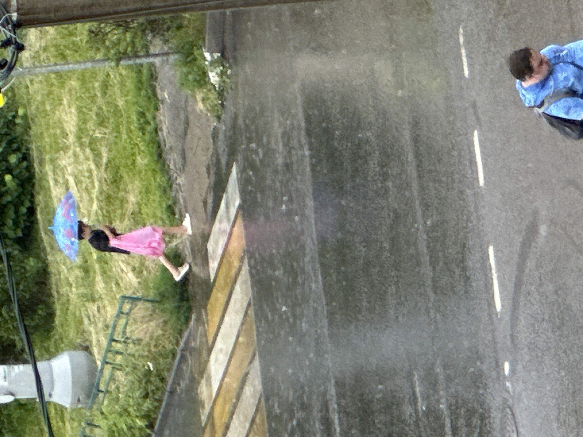 A person wearing a black shirt and a pink skirt walks under a blue umbrella on a rainy day in Podolsk, Russia. The person is on a crosswalk. The road is wet and reflects the sky. In the background, there is green grass and a metal fence. Another person wearing a blue jacket walks on the wet road.