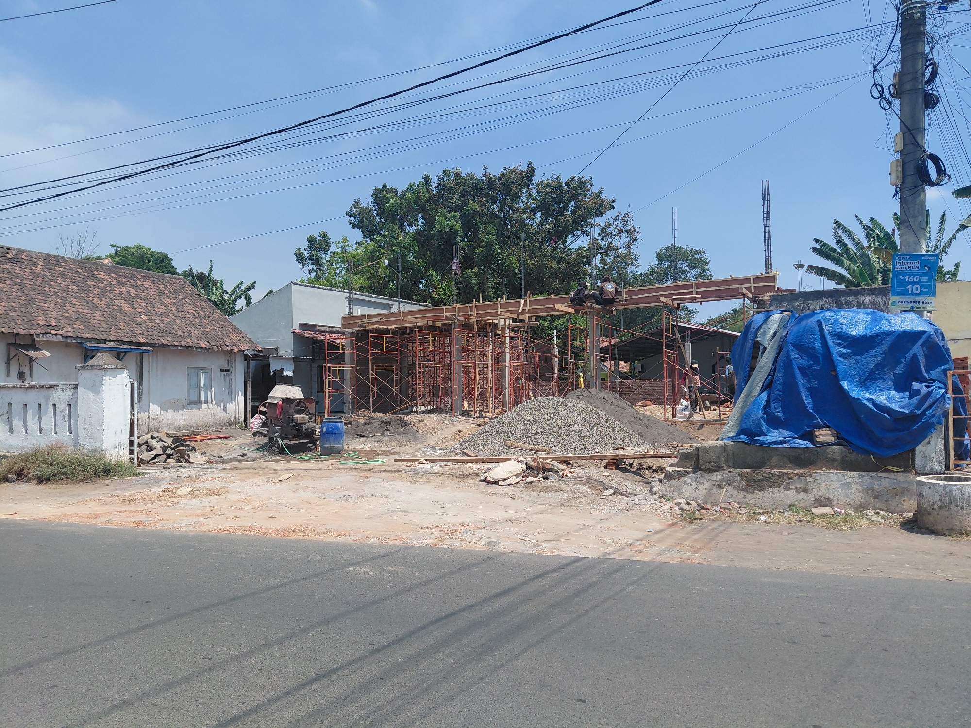 The image depicts a construction site in Pandean, Indonesia, under a clear blue sky. The site is an active construction zone with scaffolding erected around a partially built structure. Wooden beams are in place, and several workers, two of whom are visible sitting on a beam, are present. A large pile of gravel is situated in the foreground. Adjacent to the construction, an older building with a terracotta tiled roof stands to the left. Various construction materials, including a cement mixer, a blue barrel, and stacks of wood, are scattered around the site. A large blue tarp covers part of a structure on the right. Utility poles with numerous wires run across the top of the image. The scene suggests ongoing construction work on a sunny day.