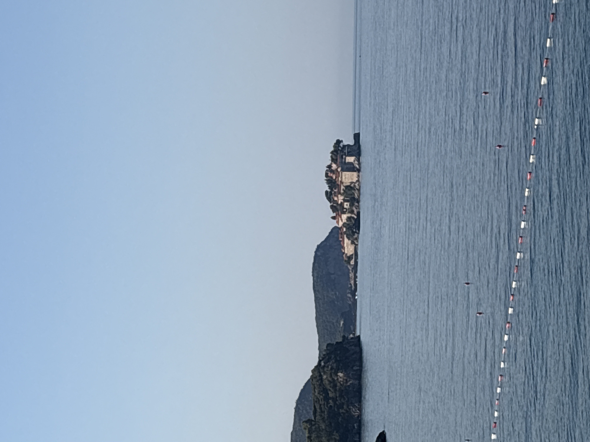 A wide shot captures the islet of Sveti Stefan, located off the coast of Kamenovo, Montenegro. The islet, a fortified village with stone buildings and red-tiled roofs, is nestled amidst lush green trees. It is connected to the mainland by a narrow strip of land. The sea, a calm expanse of blue, surrounds the islet and extends to the horizon under a clear, pale blue sky. In the foreground, a chain of white and red buoys floats on the water, marking a boundary in the sea. Two small, dark red buoys are visible further out in the water. The coastline to the left of the frame features a rugged, tree-covered hill. The overall scene depicts a serene coastal landscape on a bright, sunny day.