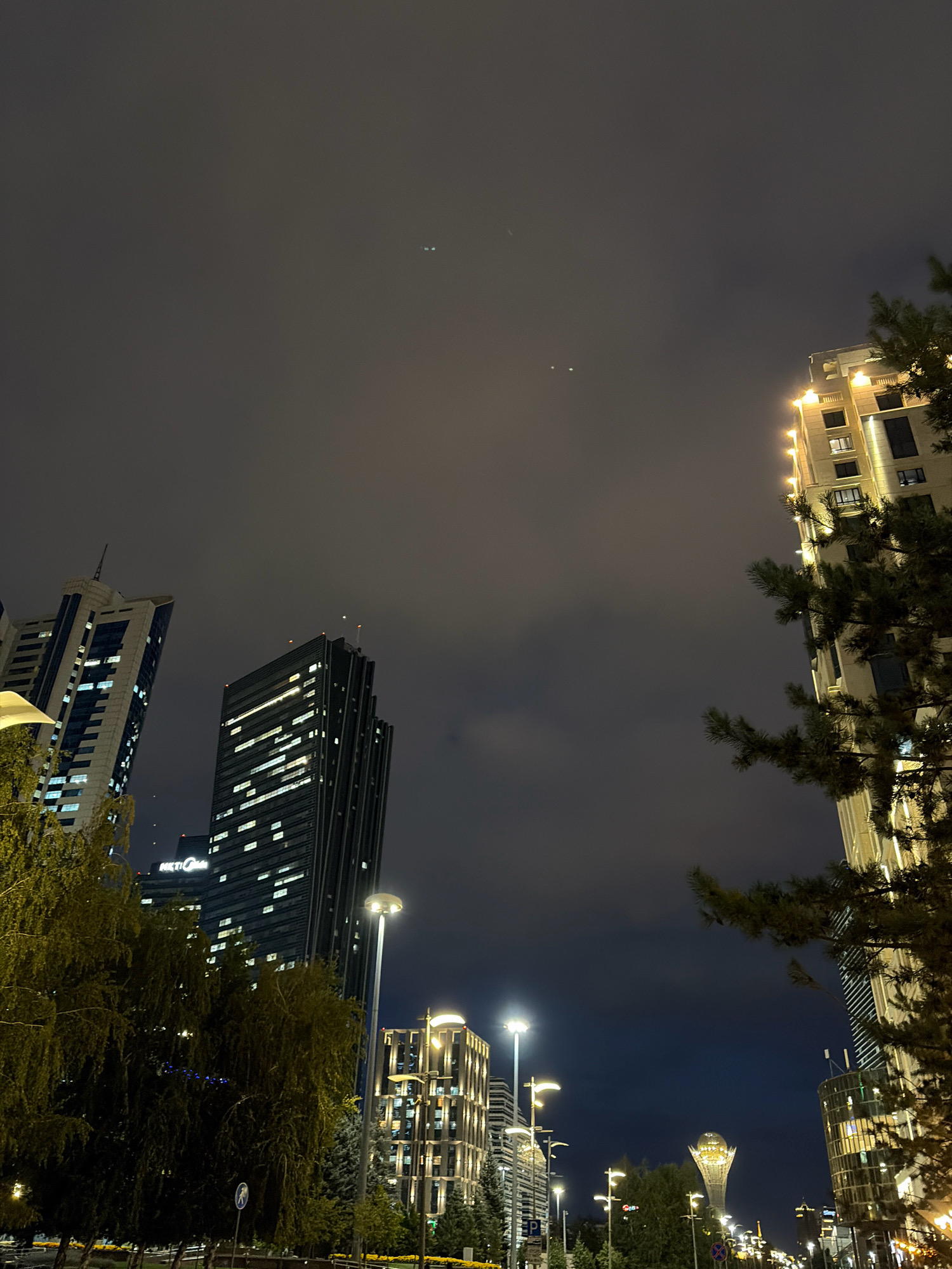 This is a nighttime photograph of the cityscape of Astana, Kazakhstan, likely from the Nur-Zhol Boulevard area, looking towards the Bayterek Tower in the distance. The sky is dark and overcast with some faint lights visible, possibly aircraft or distant stars. In the foreground and midground, several modern high-rise buildings are illuminated with interior lights. On the right, a tall, modern building with vertical golden lights is prominent, partially obscured by a large evergreen tree. To the left and center, other skyscrapers are visible with a multitude of rectangular windows lit up. A street lamp with a globe-shaped light fixture is visible in the mid-ground, casting a yellow glow. In the far distance, the distinctive silhouette of the Bayterek Tower is discernible, illuminated against the night sky. Trees and some street signage are also present in the lower portions of the image. The overall impression is an urban environment at night, characterized by modern architecture and artificial illumination.