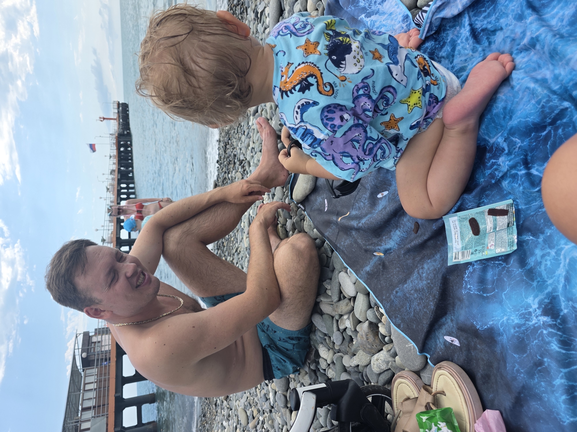 The image depicts a sunny day at a pebble beach in Sochi, Russia. A man is sitting on a blue blanket spread over the pebbles, facing left and smiling. He is shirtless, wearing turquoise swim shorts, and a gold chain. A toddler, seen from behind, is also sitting on the blanket, wearing a light blue t-shirt with various sea creatures printed on it. The toddler's legs and feet are visible, and they appear to be playing with something on the blanket. A stroller is partially visible to the left of the man. In the background, a pier extends into the sea, with a building on stilts and a red lighthouse at the end. The Russian flag flies from a flagpole on the pier. The sea is calm, and the sky is partly cloudy.