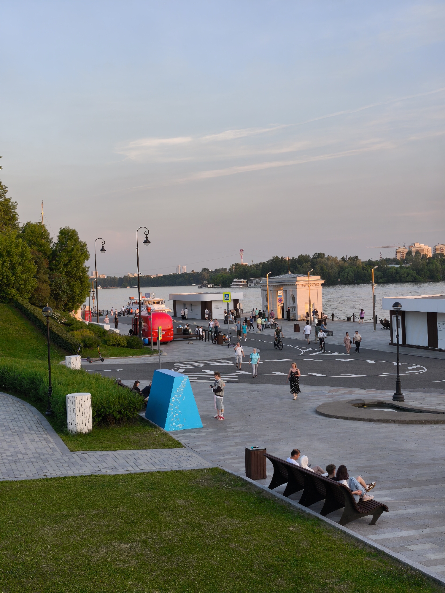 This image depicts a late afternoon scene at the North River Station (Severny Rechnoy Vokzal) embankment and park in Moscow, Russia.
The sky is a pale blue with faint, wispy clouds. In the foreground, a verdant grassy slope on the left transitions to a grey paved walkway. A dark brown wooden bench with a backrest is positioned on the right side of the paved area, occupied by three individuals; two adults are seated with a small child or infant partially obscured between them. Further left on the paved area, a white cylindrical bollard with decorative cutouts stands next to a patch of green bushes. Adjacent to the bushes, a bright blue triangular prism sculpture with white graphical patterns on its surface is visible. A person in a grey hoodie and shorts stands near this sculpture, appearing to interact with a handheld device.
The midground features a wide, paved promenade alongside the Moscow River. White painted markings, including arrows and curved lines, delineate paths on the asphalt. Numerous pedestrians, dressed in casual attire, are walking in various directions across the promenade. Two individuals are seen riding bicycles. A red double-decker vehicle, likely a tour bus, is parked near the water's edge on the left. Several dark-colored street lamps with decorative scrollwork top the poles. Near the river, a classical white building with columns bears the inscription "ПРИЧАЛ №5" (Pier No. 5). People are congregated around this building and along the pier. To the right of the classical building, a modern white building with a flat roof stands. A yellow pedestrian crossing sign is visible near the classical building.
The background shows the calm waters of the Moscow River, where several boats and ships are either docked or navigating. The opposite bank is densely covered with green trees. In the far distance, various urban buildings and structures, including construction cranes, are visible, indicating a developed cityscape.