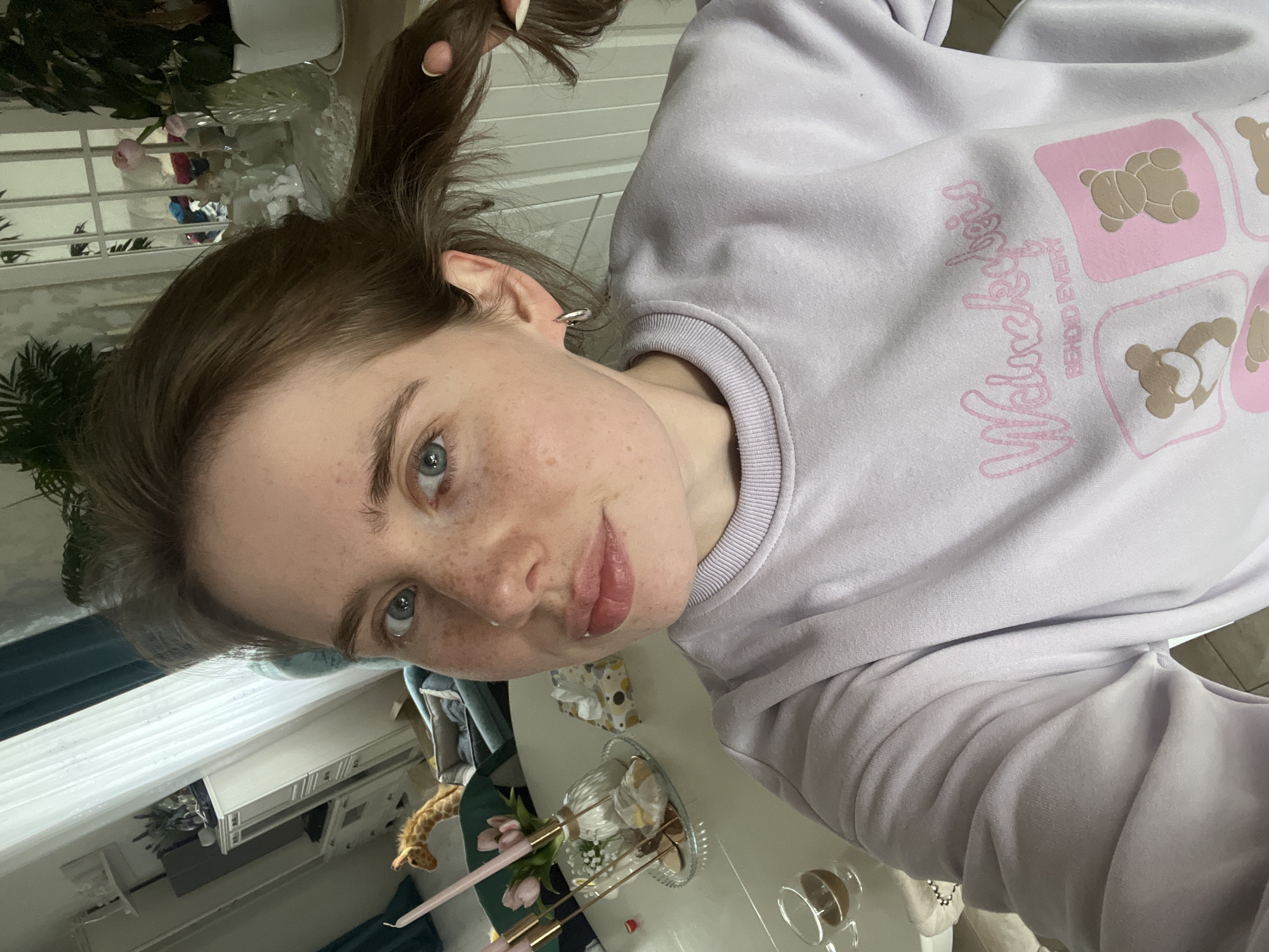 A young woman with light skin, freckles, and blue eyes is facing the camera. Her hair is pulled back in a ponytail. She is wearing a light purple sweatshirt with teddy bear designs and pink lettering that reads "Volochkova Behind Every." She appears to be indoors, possibly in a room with a window, a table, and some plants. On the table, there is a decorative dish with pastries and flowers. There are also some candles and a glass on the table. A plush giraffe toy is situated in the background near the table. The location is specified as the city of Chaikovsky, Russia.