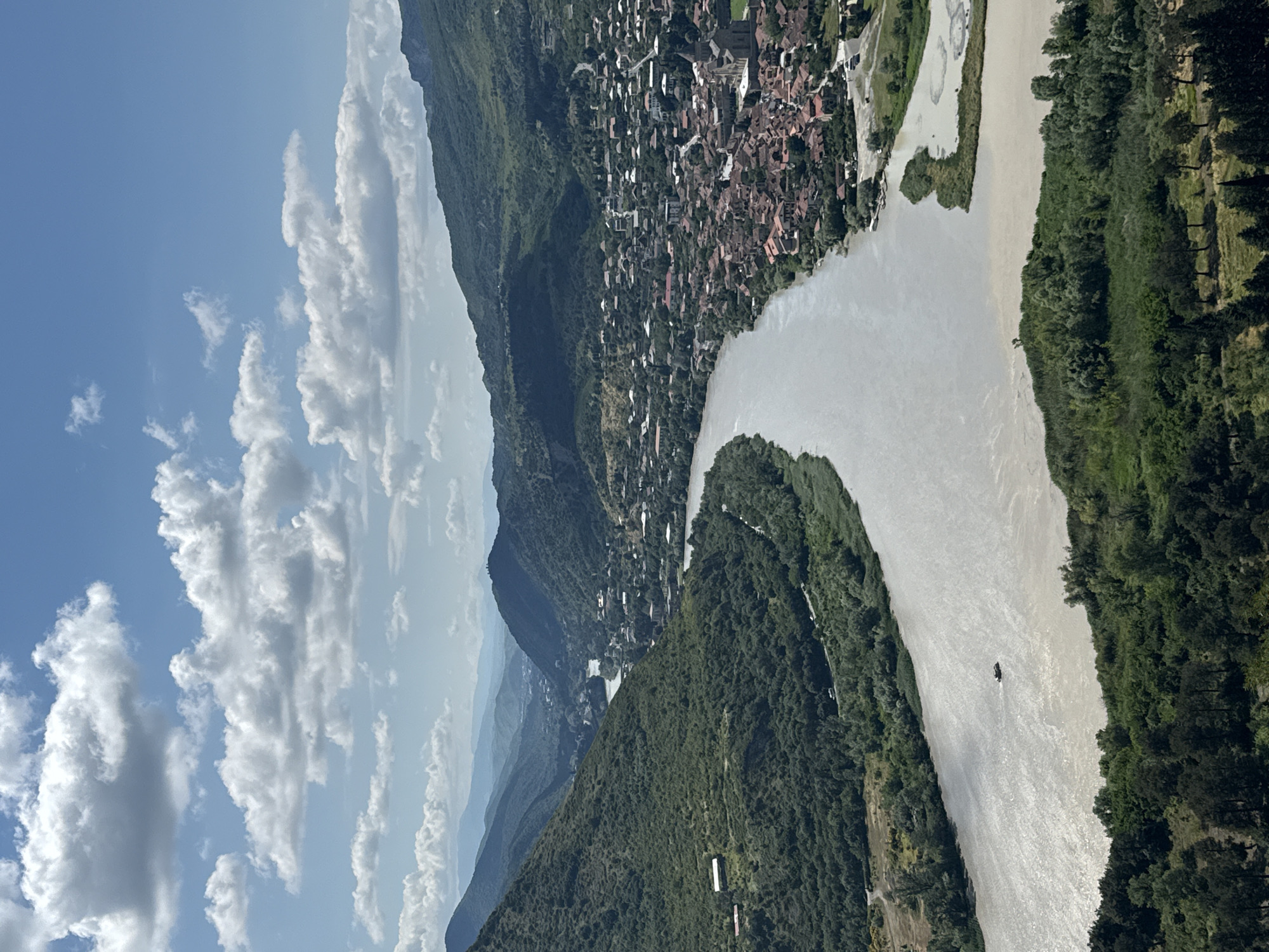 This is an aerial view of a city nestled within a valley, with a wide river flowing through it. The city, identified as Mtskheta, is located in Mukhatgverdi, Georgia. The buildings are characterized by red-tiled roofs, clustered together along the riverbanks and extending up the hillsides. A prominent stone church with a dark roof is visible within the city. The river itself appears muddy and wide, with a small boat visible in the water. The surrounding landscape is dominated by lush green mountains and hills, covered in dense vegetation. The sky is a bright blue with scattered white clouds, suggesting a clear and sunny day. The overall scene depicts a historic city integrated with its natural mountainous environment.