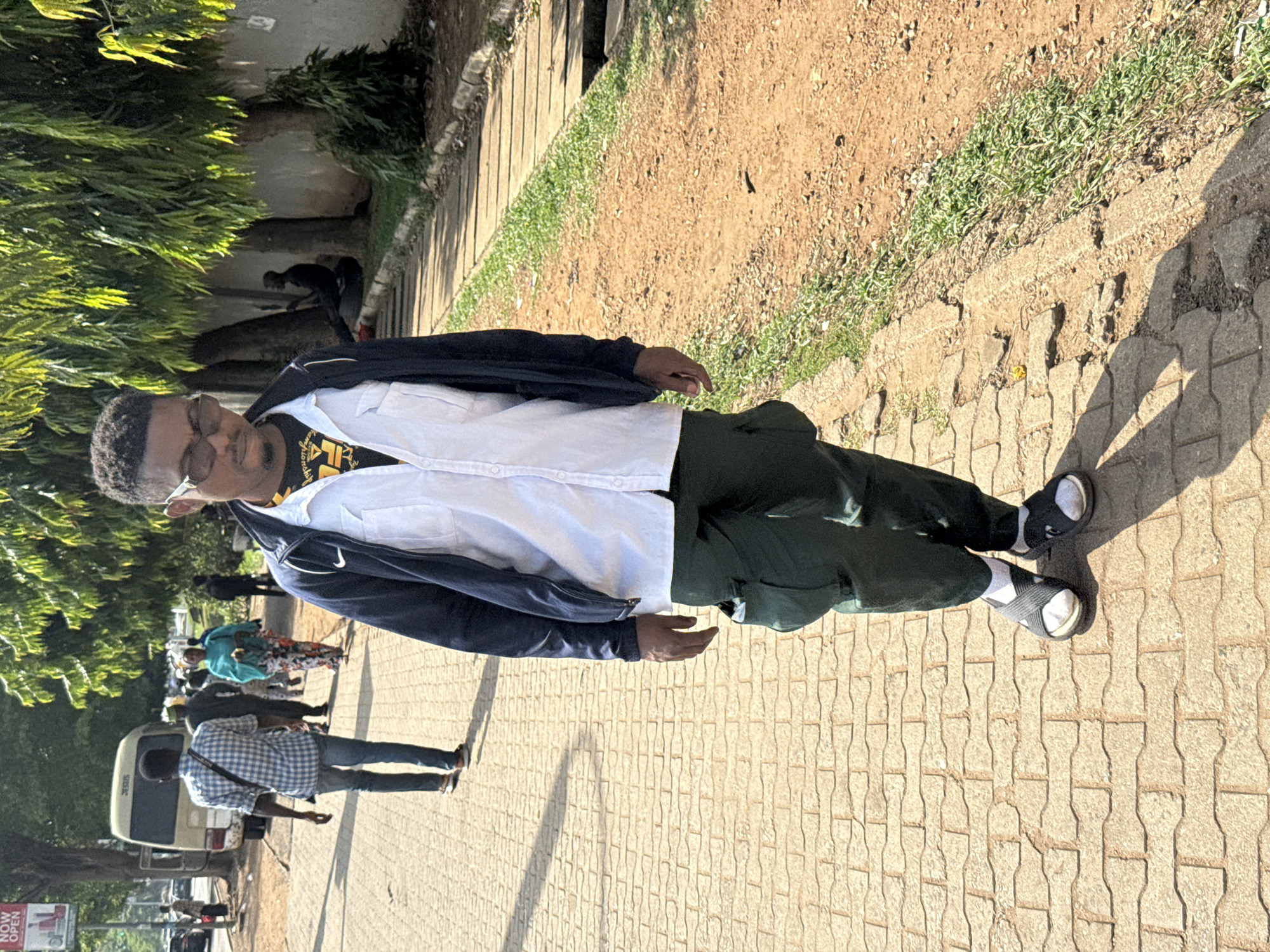 A man wearing glasses, a white button-down shirt, a dark-colored jacket, dark pants, and sandals is standing on a sidewalk in a sunlit area. The sidewalk is paved with light-colored interlocking pavers. To the man's right is a dirt area with some grass and a raised concrete or stone border. To his left, there are trees and other people, including someone near a parked vehicle. A sign is visible in the lower left corner. The location appears to be in a populated area within Abuja, Nigeria.