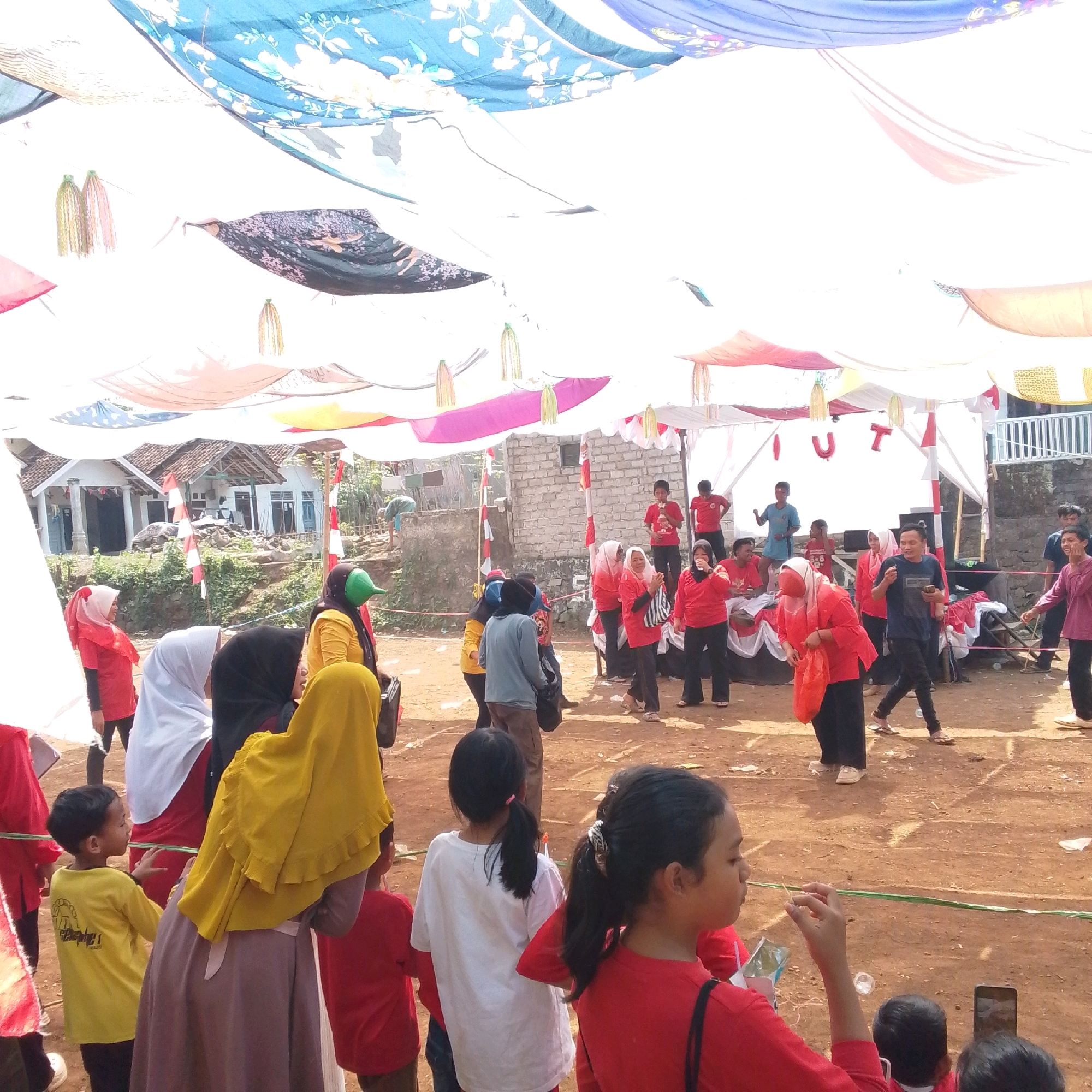 The image depicts an outdoor event, possibly a community gathering or celebration, in Cilaku, Indonesia. A large canopy made of colorful fabrics, some with floral patterns and others plain, is set up to provide shade. Red and white Indonesian flags are visible, suggesting a national holiday or celebration. Numerous people, including children and adults, are present, many wearing red shirts. Some are engaged in activities, while others are observing. In the background, traditional Indonesian houses with tiled roofs are visible. The ground appears to be dirt, indicating a rural or semi-rural setting. The overall atmosphere seems festive and communal.