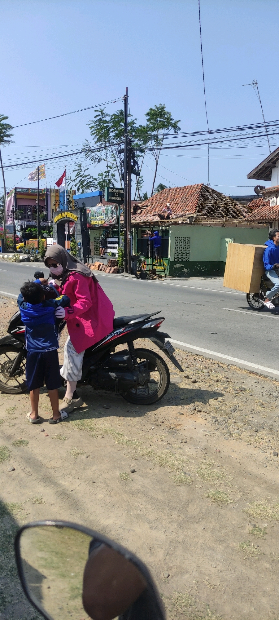In the city of Purwodadi, Indonesia, a woman wearing a pink jacket, grey hijab, white and grey striped pants, and a surgical mask is interacting with a young boy wearing a blue jacket and navy shorts. They are standing next to a black motorcycle parked on the side of a paved road. The woman appears to be holding onto the boy. The boy is facing the woman and appears to be reaching out. In the background, several men are working on the roof of a green building with a red tiled roof. One man is on a ladder, while others are on the roof. Further in the background, there are other buildings, flags, and utility poles with wires. The scene is illuminated by daylight.