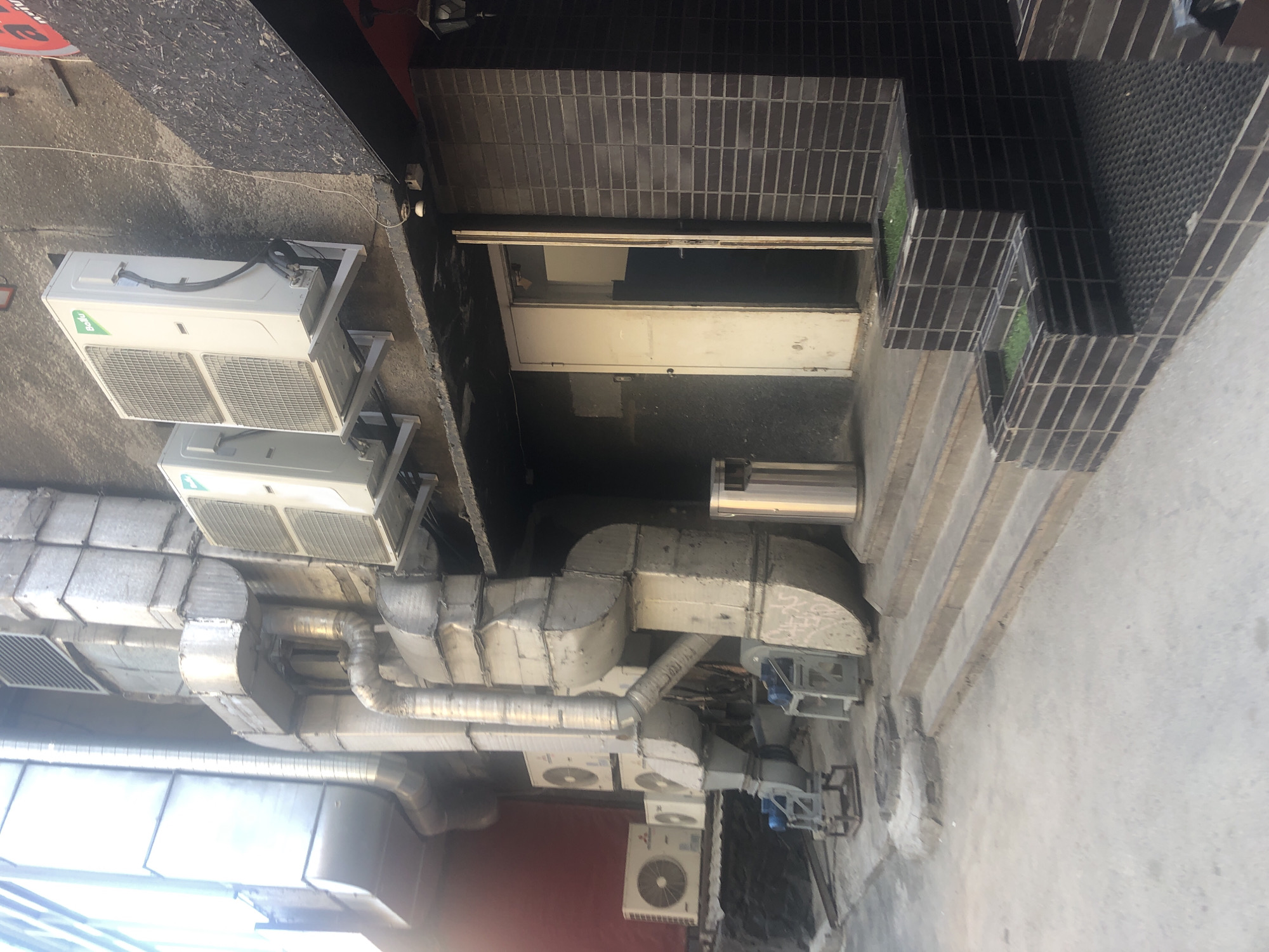 A wide outdoor shot captures a section of a building's exterior, featuring air conditioning units, ventilation ducts, a doorway, and a set of stairs. In the foreground, a concrete walkway leads to a set of stone steps. To the left, several outdoor air conditioning units are visible, some mounted on the wall, others on the ground with visible fans. Large, silver ventilation ducts are prominently displayed, running vertically and horizontally along the building's facade. A closed doorway, partially open, is situated to the right of the ventilation system, revealing a dark interior. The building's wall is textured, with some areas appearing to be rough concrete and others covered in dark rectangular tiles. A stainless steel trash can is positioned near the steps. The scene is illuminated by daylight, casting shadows on the ground.