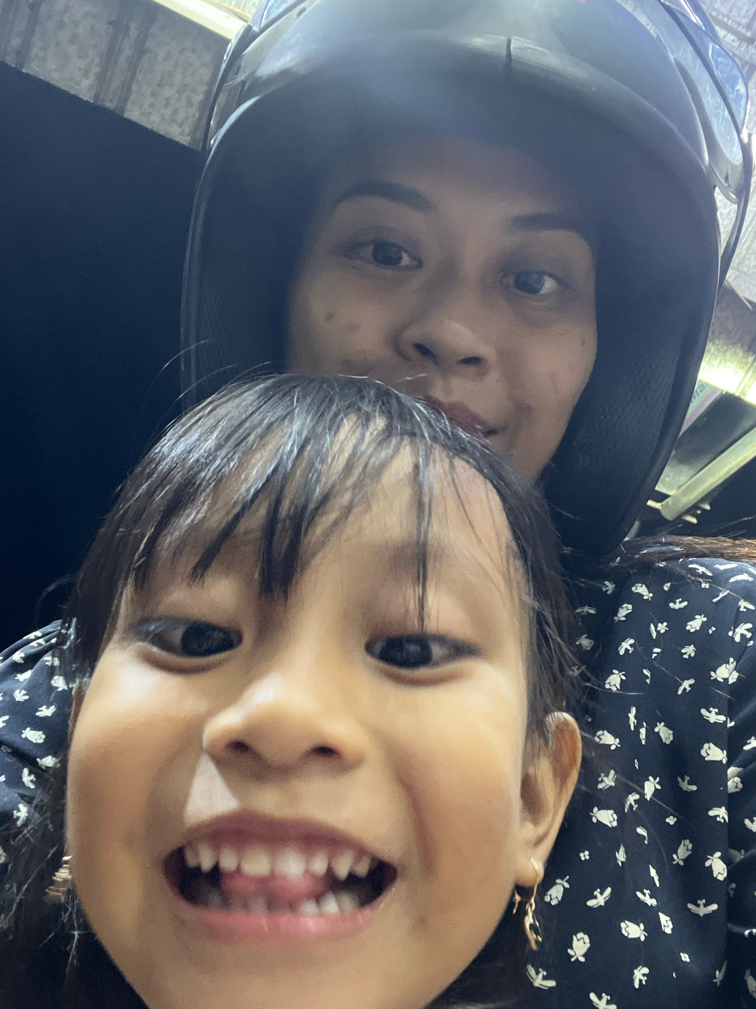 A medium close-up selfie shows two people, an adult and a child, positioned in front of a dark, possibly outdoor, setting. The adult is in the background, wearing a black helmet. The child is in the foreground, their face filling most of the frame. The child is looking directly at the camera with a wide smile, revealing their teeth and tongue. They have dark hair that is slightly messy, falling forward onto their forehead and cheeks. The child is wearing a dark-colored shirt with a small white floral pattern. An earring is visible on their right ear. The adult's face is partially obscured by the helmet and their proximity to the child. The image appears to have been taken on a street or thoroughfare in Kecamatan Pati, Indonesia, given the context. The lighting suggests it might be daytime, possibly late afternoon or early evening.