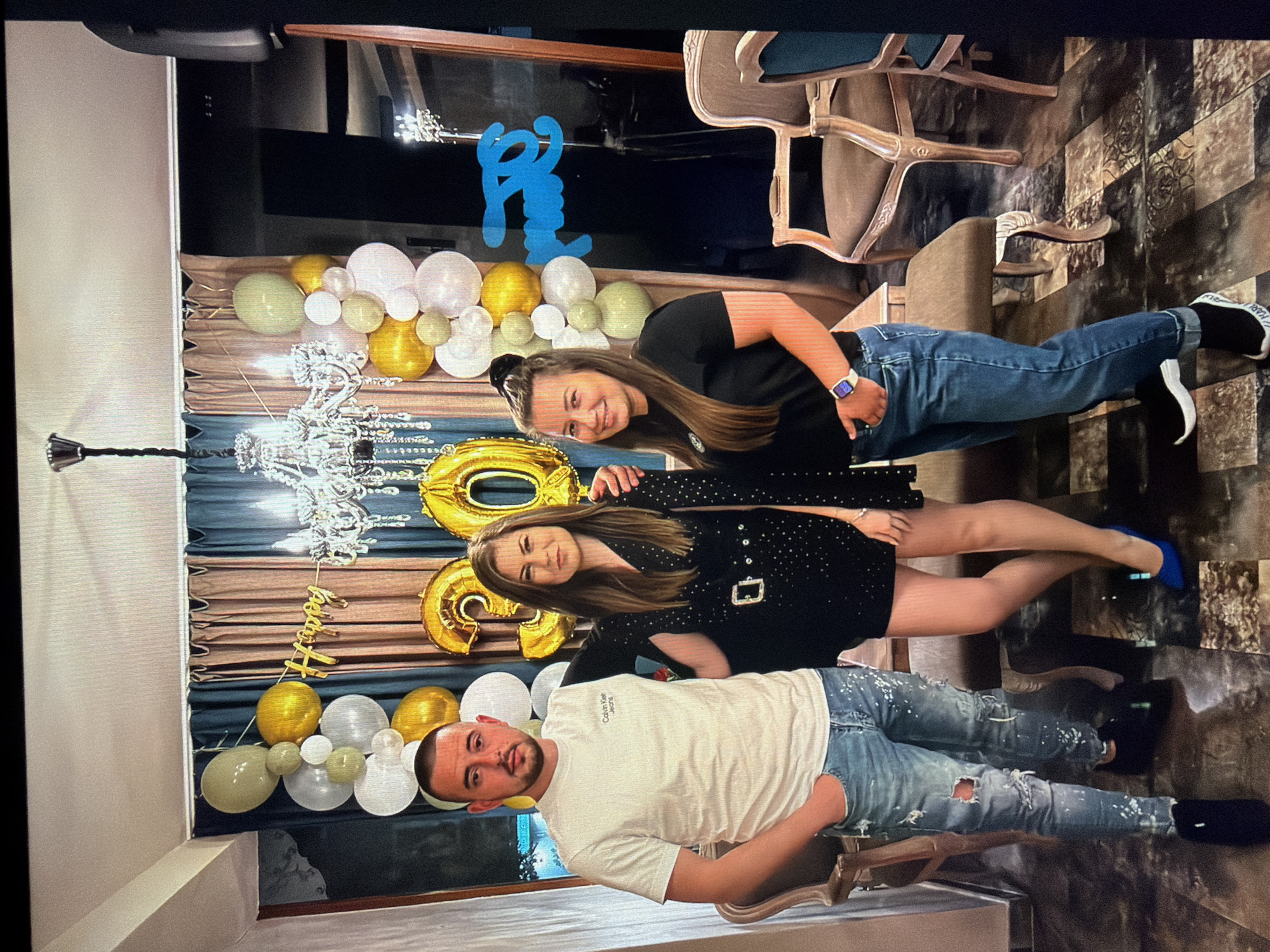 The image shows three individuals posing for a photo indoors. A man wearing a white t-shirt and distressed jeans stands in front, with two women behind him. One woman wears a black t-shirt and jeans, and the other wears a black, short dress with a belt. The background includes a balloon display with "Happy" and the number "30" in golden balloons. There is also a chandelier and draped fabric. The flooring is tiled with a brown, patterned design. Chairs are also visible in the background. The location is likely in Burgas, Bulgaria.