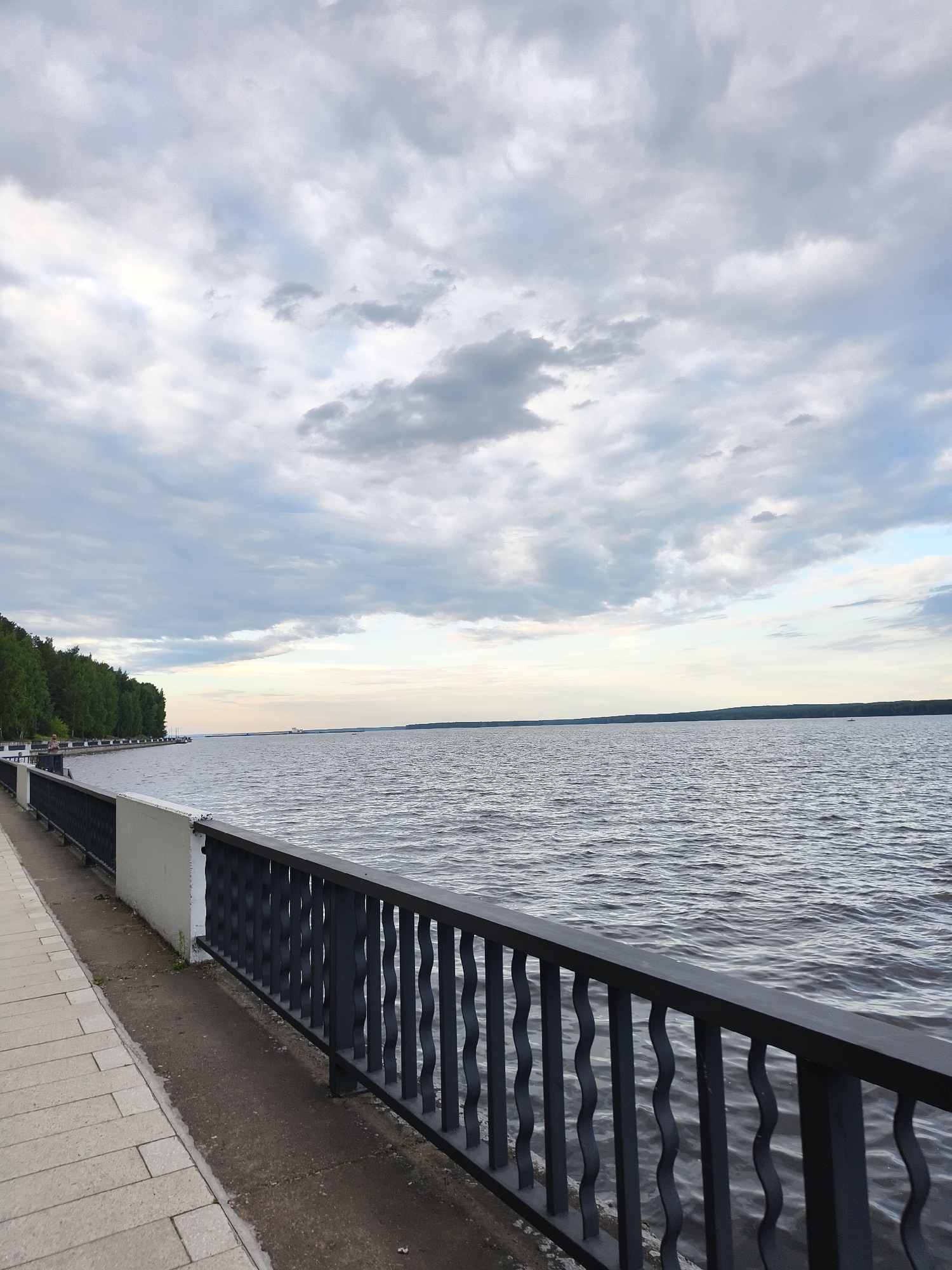 The image depicts a waterfront promenade in Chaikovsky, Russia, with a broad river occupying the majority of the frame. A dark metal railing runs along the edge of the promenade, separating it from the river. The promenade itself is paved with light-colored rectangular tiles. To the left, a dense line of green trees borders the embankment. The sky is overcast with scattered clouds, casting a muted light over the scene. In the distance on the river, a large barge or ship is visible. A single person can be seen standing on the promenade, looking out at the water. The water's surface is rippled, indicating a gentle breeze. The overall atmosphere is calm and serene.