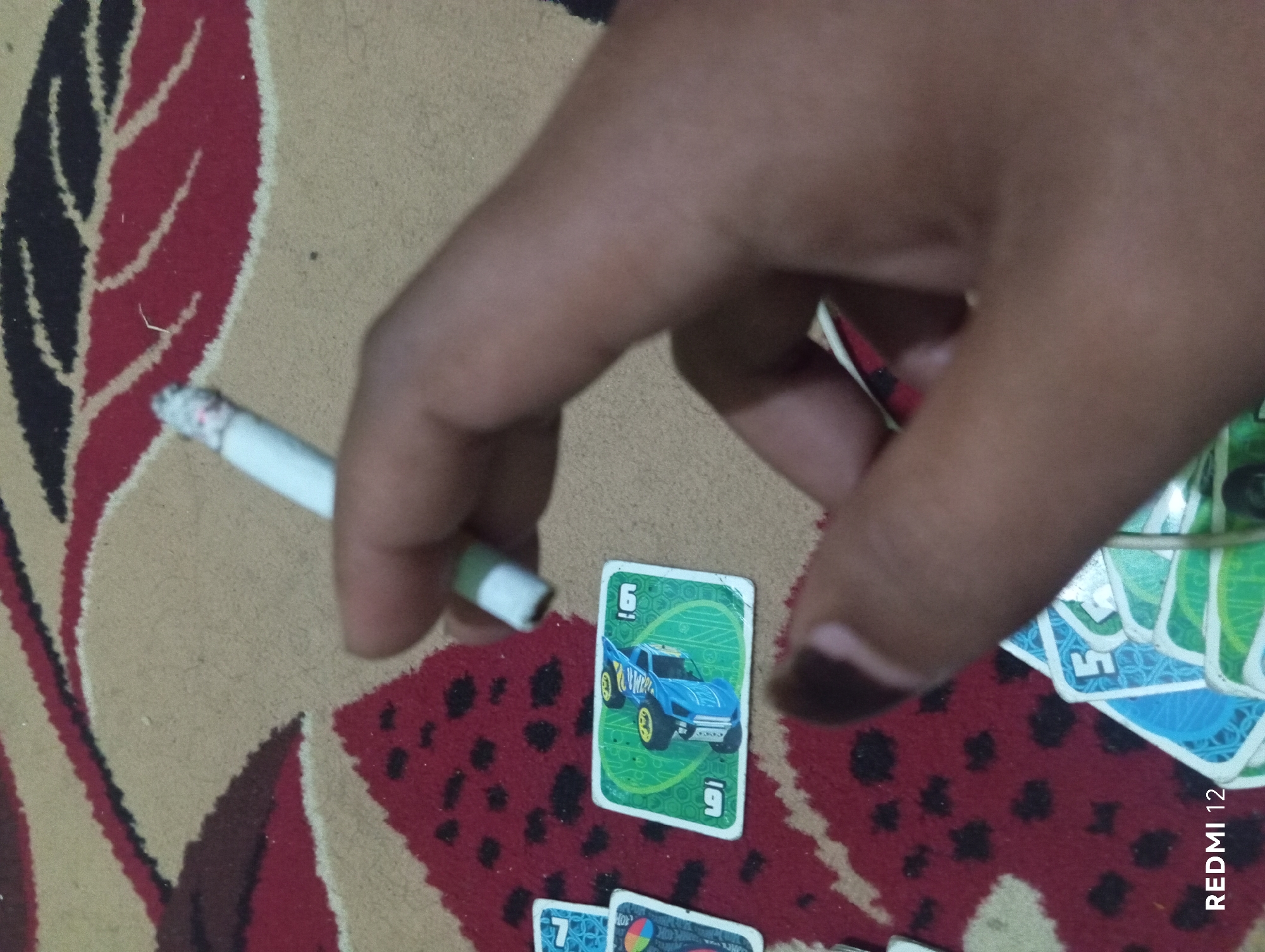 A close-up, overhead shot shows a person's hand holding a lit cigarette over a pile of playing cards on a red and tan patterned carpet. One card, featuring a blue toy car and the number "9" in the corner, is clearly visible. The hand's skin tone is brown. The background consists of the carpet with a repeating leaf-like pattern in red, black, and tan. The overall scene is informal and appears to be indoors. The image is tagged with "REDMI 12," indicating the phone model used for capture.
